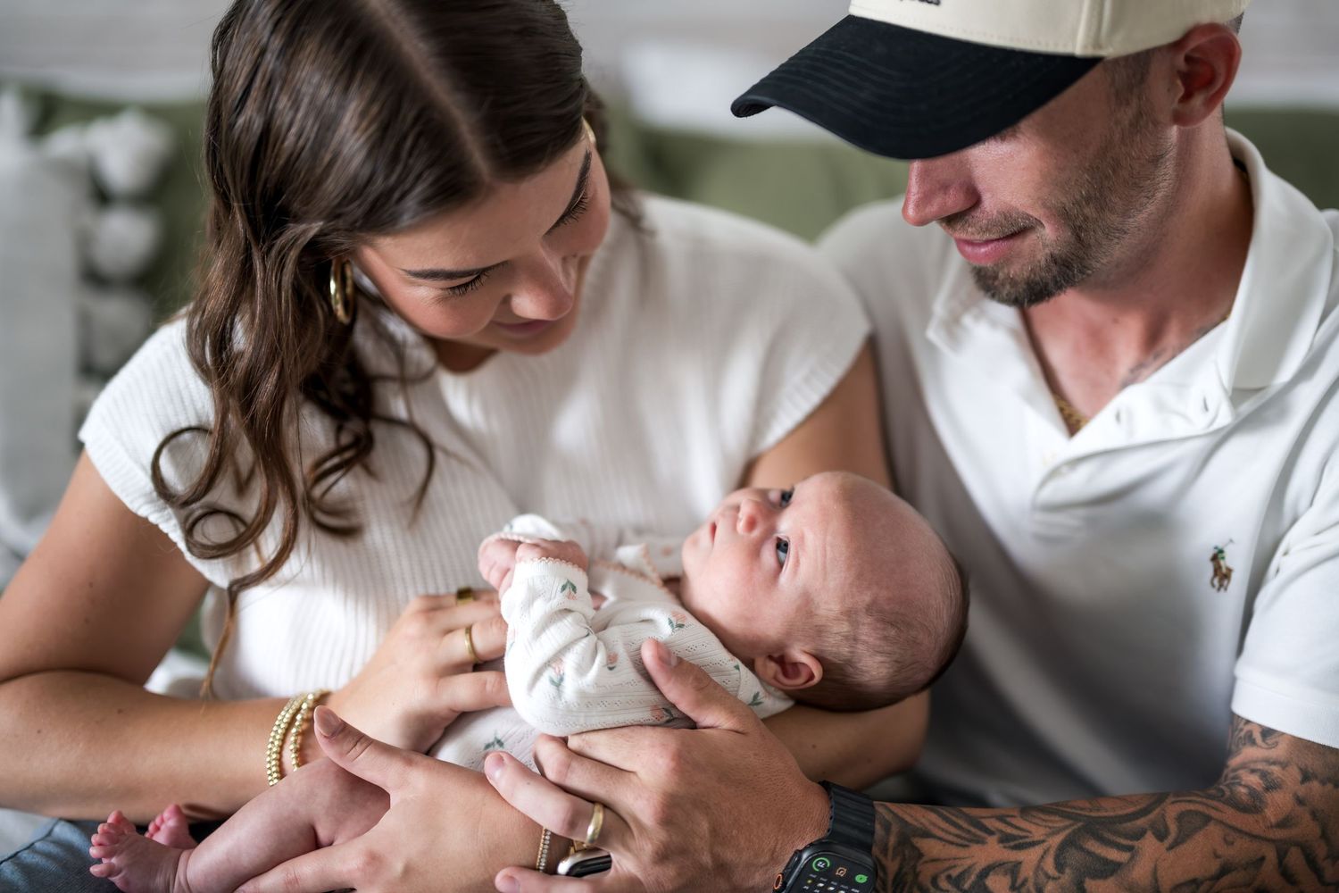 A couple lovingly gazes at their newborn baby, with the tattooed father wearing a navy cap and white polo shirt.