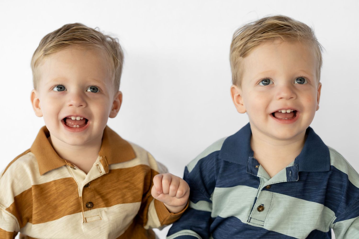 Two smiling toddler twin boys with blonde hair wearing striped polo shirts posed against a white background.