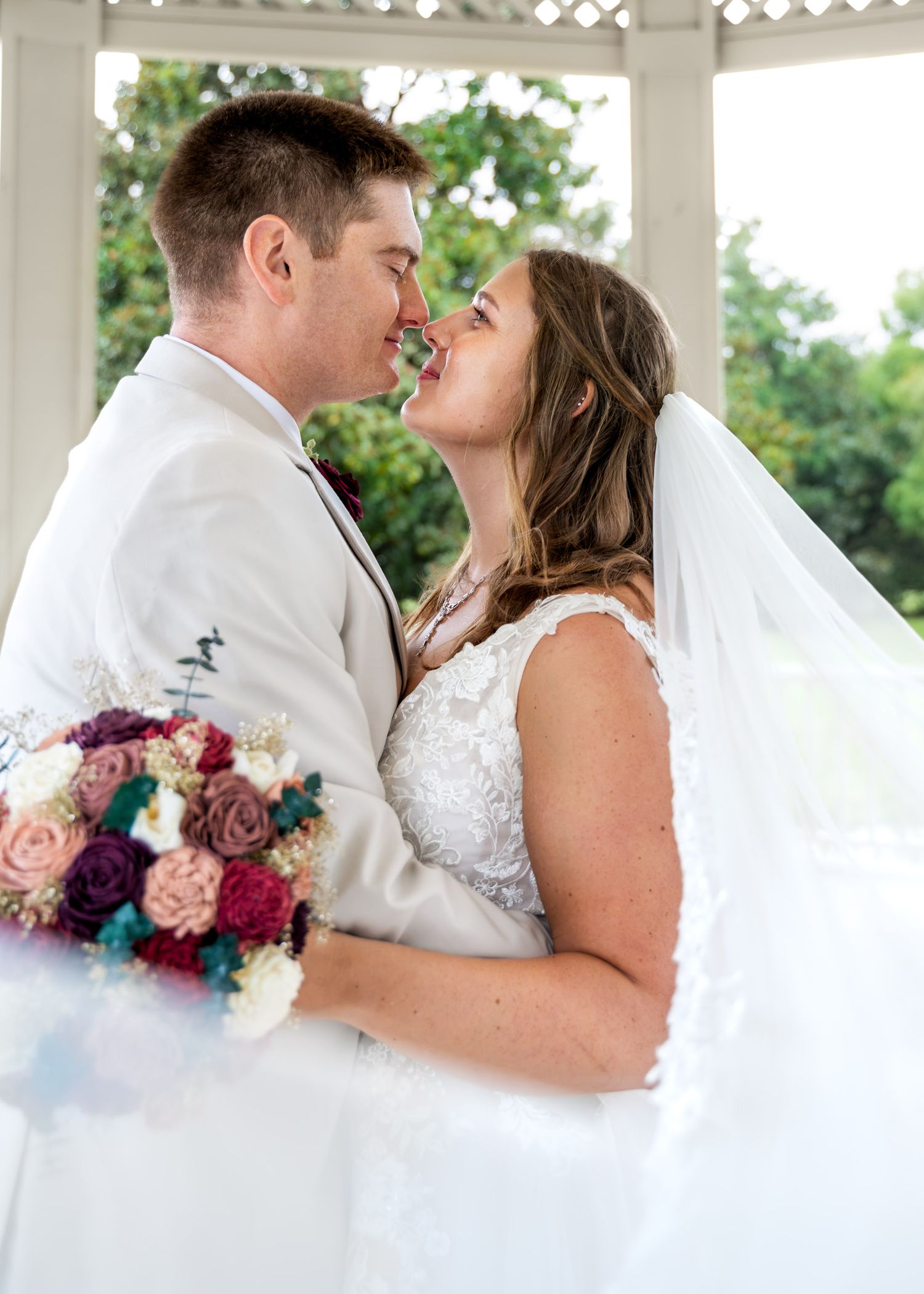 Hampton Park. Bride and groom nearly kissing in white gazebo, bride holding colorful burgundy and blush bouquet.
