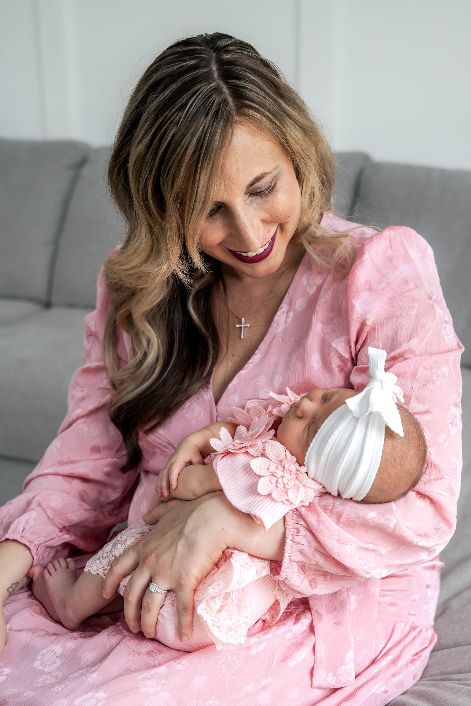 Smiling blonde mother in pink floral dress tenderly holds her newborn baby girl with bow headband.