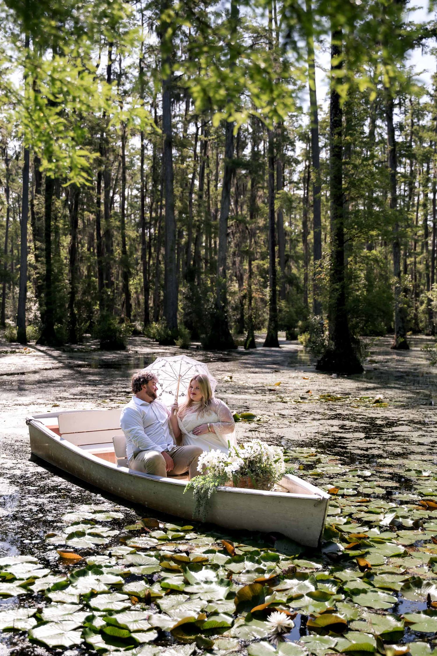 Couple sitting in a canoe surrounded by lily pads in a serene forest swamp, holding a parasol.