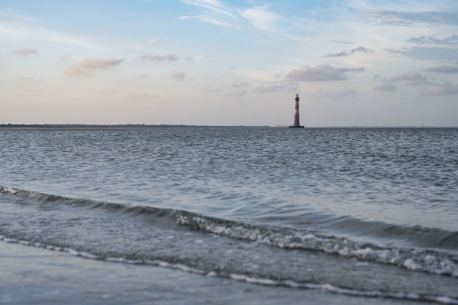 A distant lighthouse stands in calm ocean waters with gentle waves on a cloudy beach at dusk.
