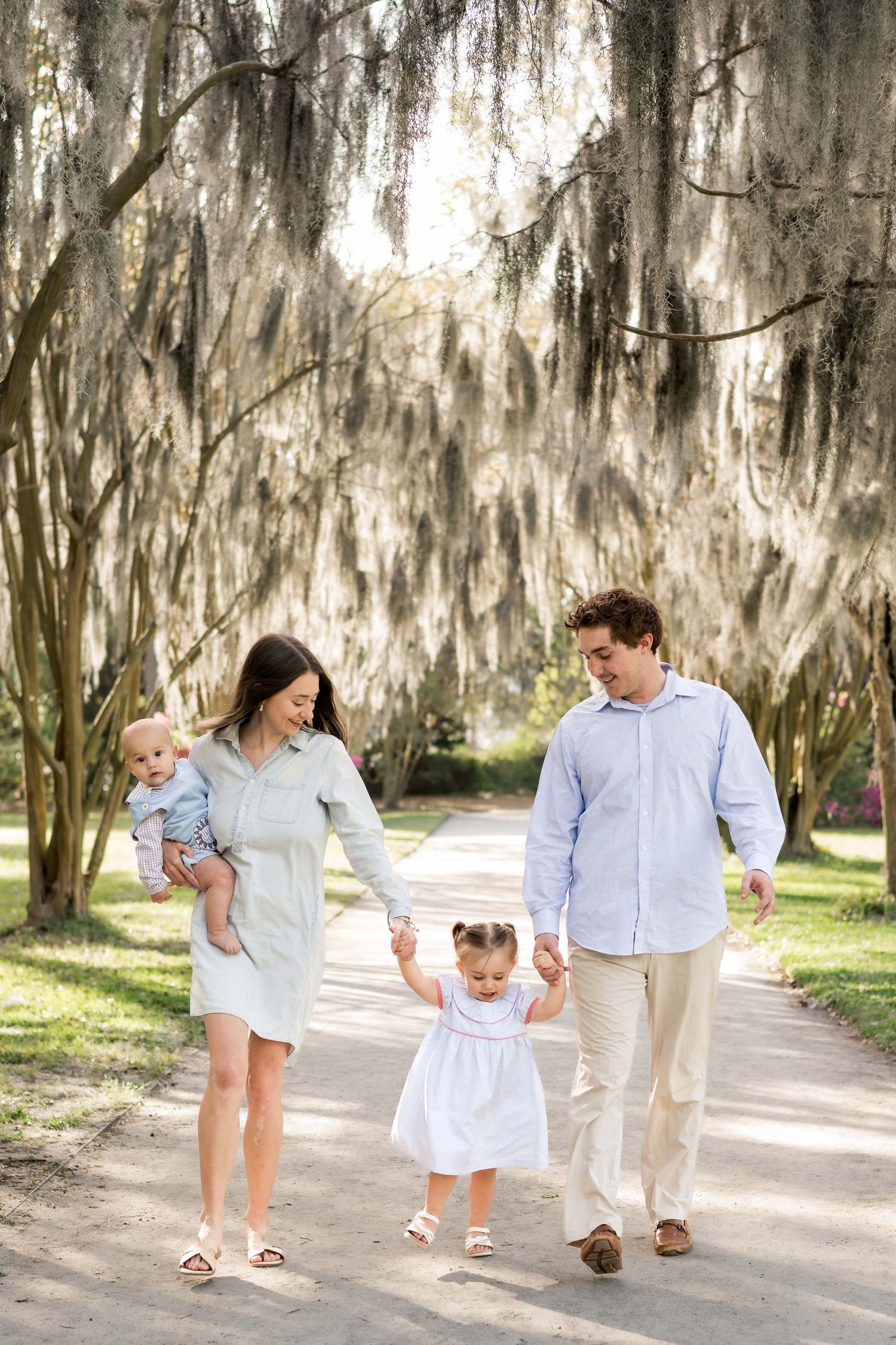 Family of four walking on a tree-lined path with Spanish moss, parents holding toddler's hands while mom carries baby.