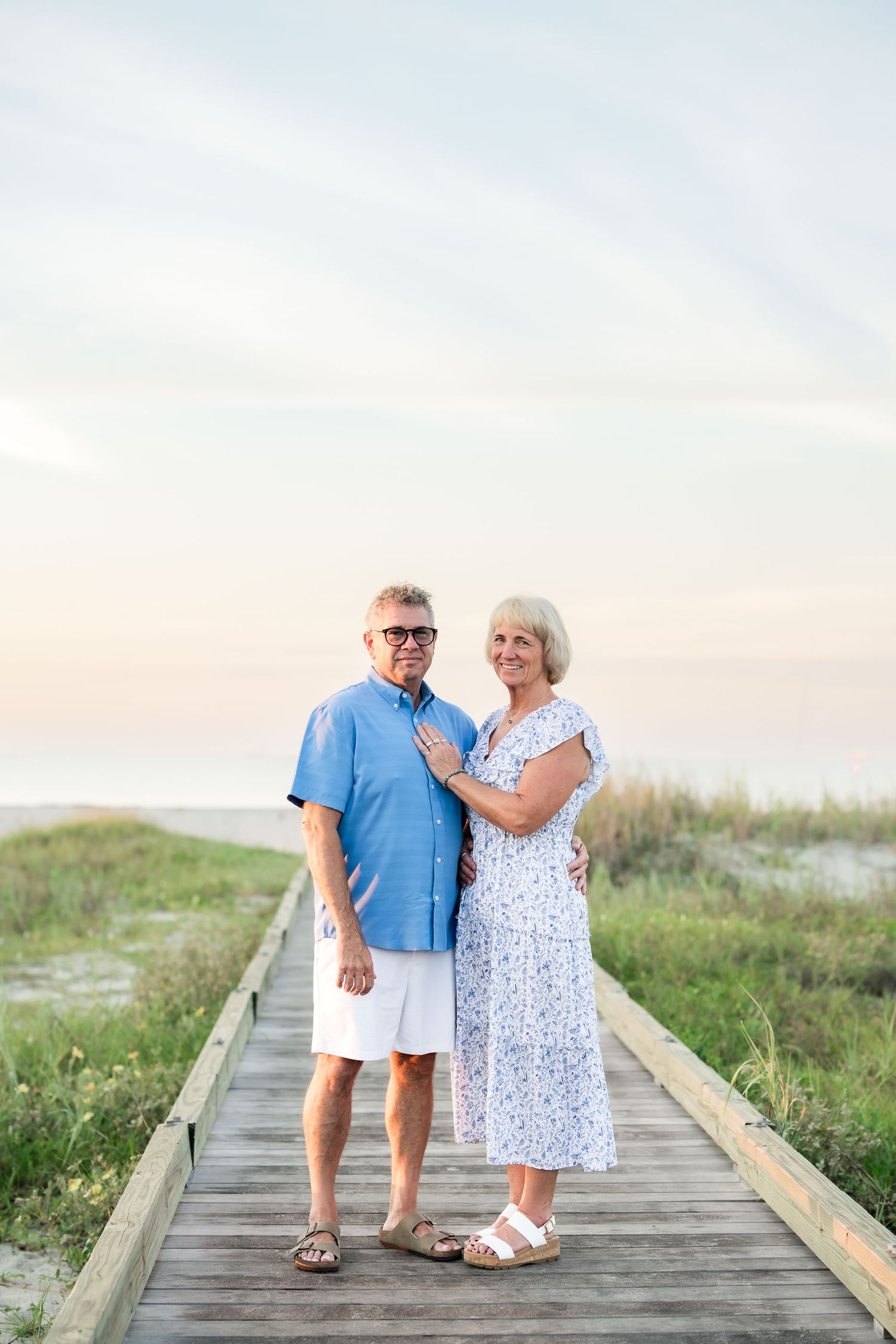 A smiling older couple poses on a beach boardwalk at sunset, dressed in blue and white summer attire.