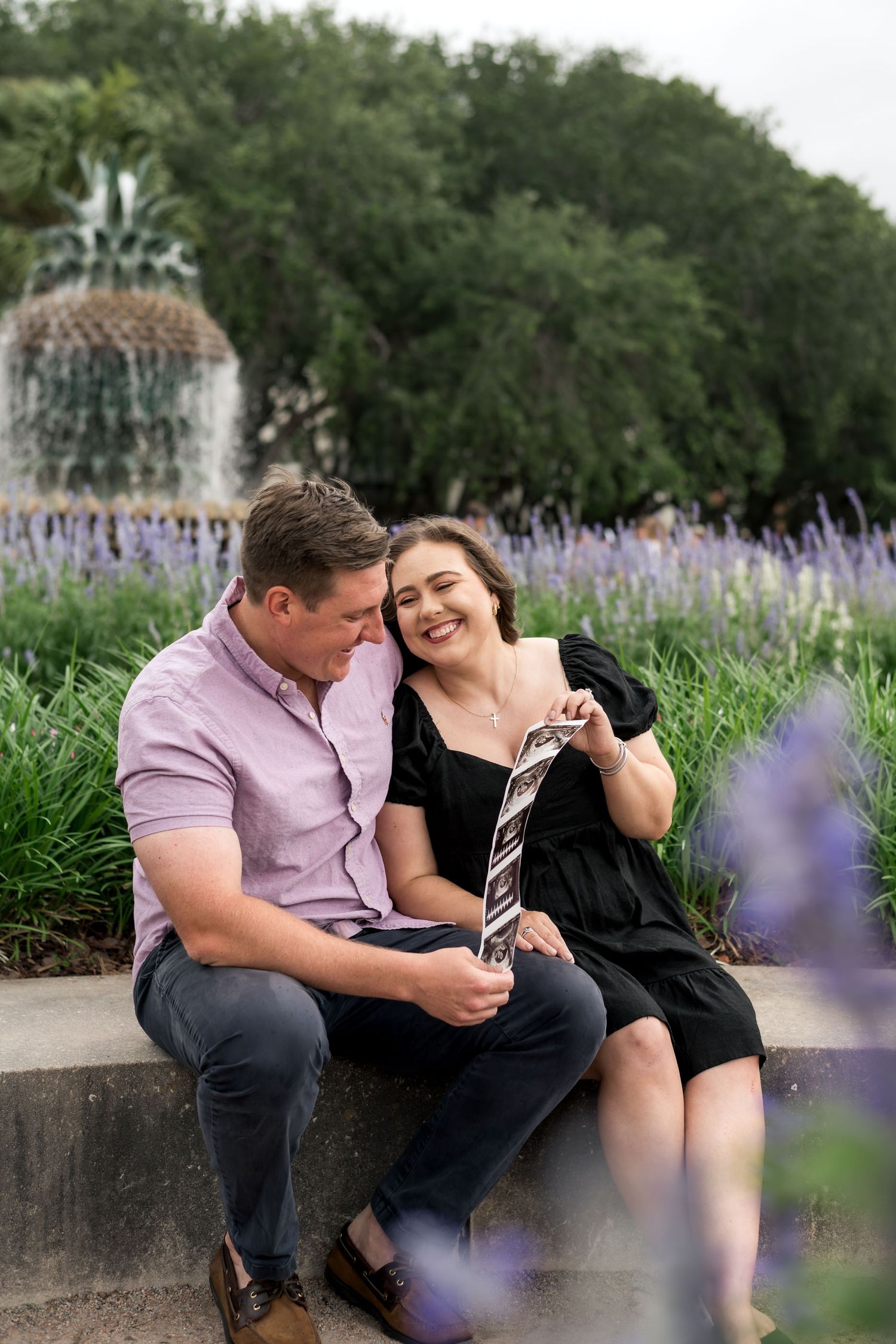 Pineapple Fountain. A happy couple sits by a fountain surrounded by purple flowers, sharing a sweet intimate moment together.