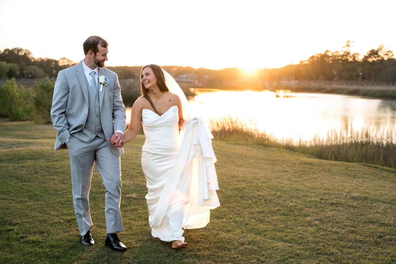 Bride and groom walk hand in hand at golden hour beside a serene lake on their wedding day.