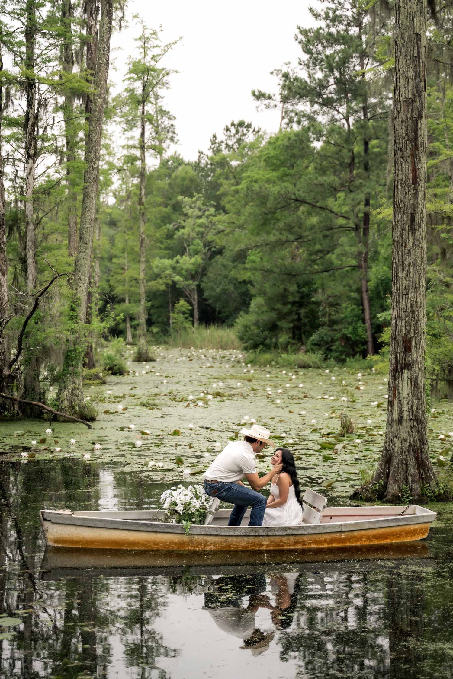 Couple sharing a romantic moment in a wooden rowboat on a serene cypress swamp, surrounded by lily pads and lush greenery.