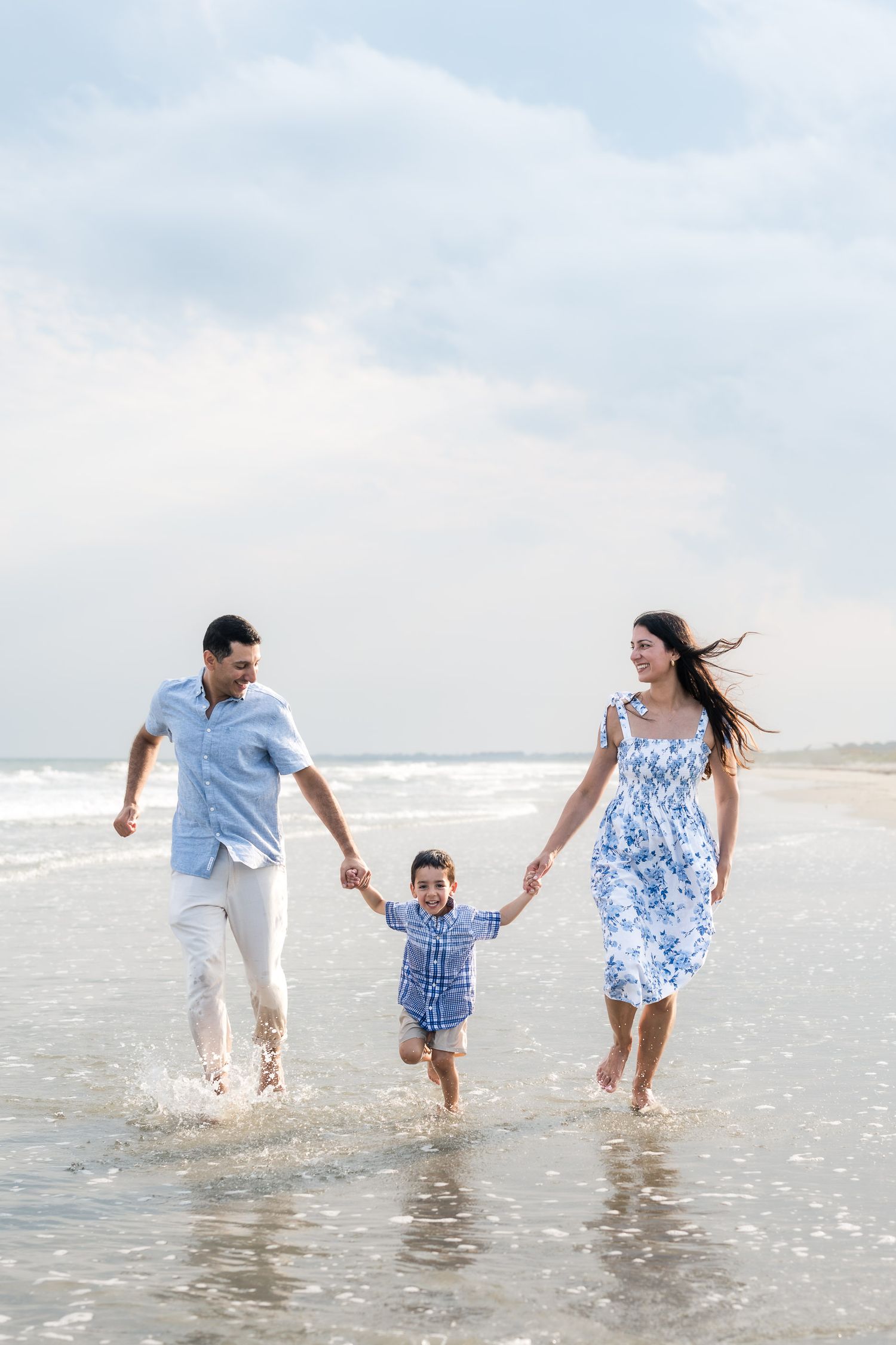 Joyful family of three holding hands and splashing through ocean waves on a beautiful beach day.