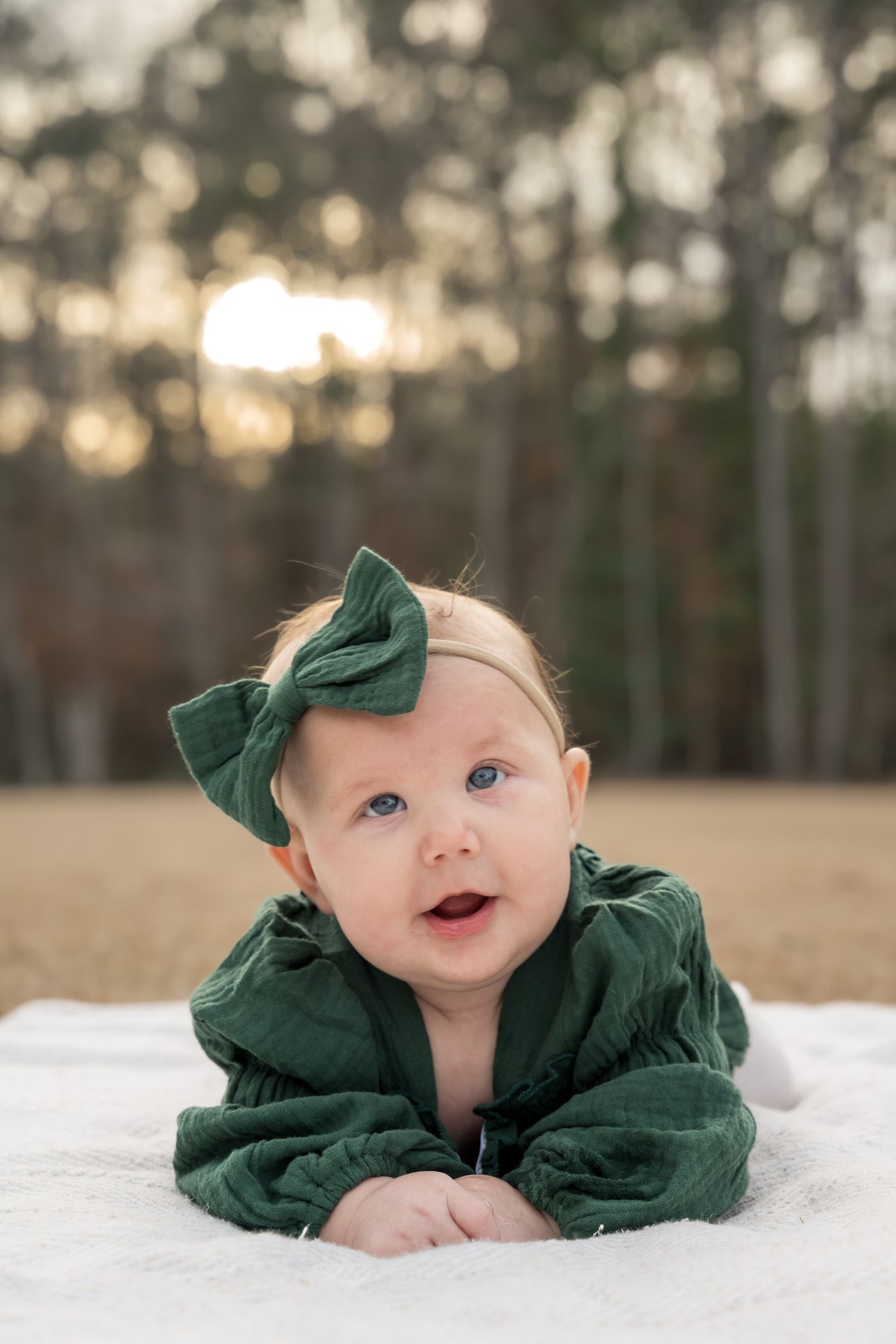 Adorable baby girl in green outfit and bow headband doing tummy time on white blanket outdoors.