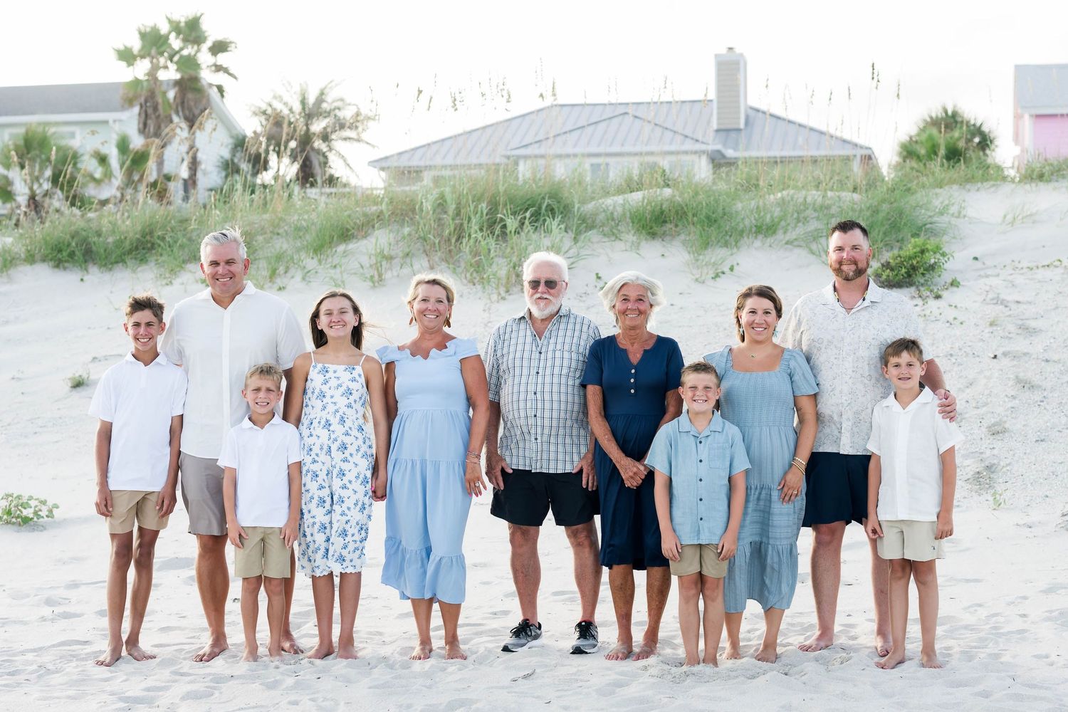 Multi-generational family portrait on sandy beach, smiling group in coordinated white and blue outfits with ocean house backdrop.