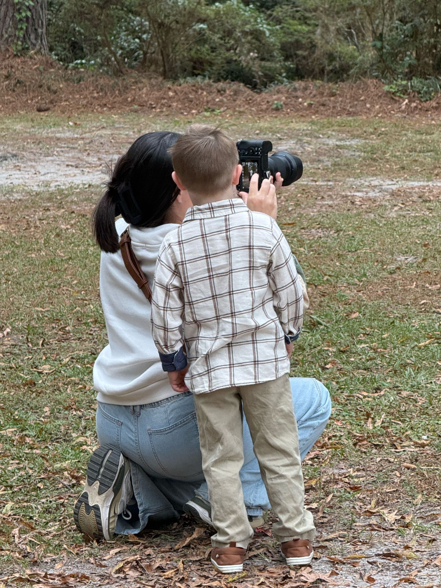 A photographer in casual attire crouches low to capture a shot outdoors in a wooded area.