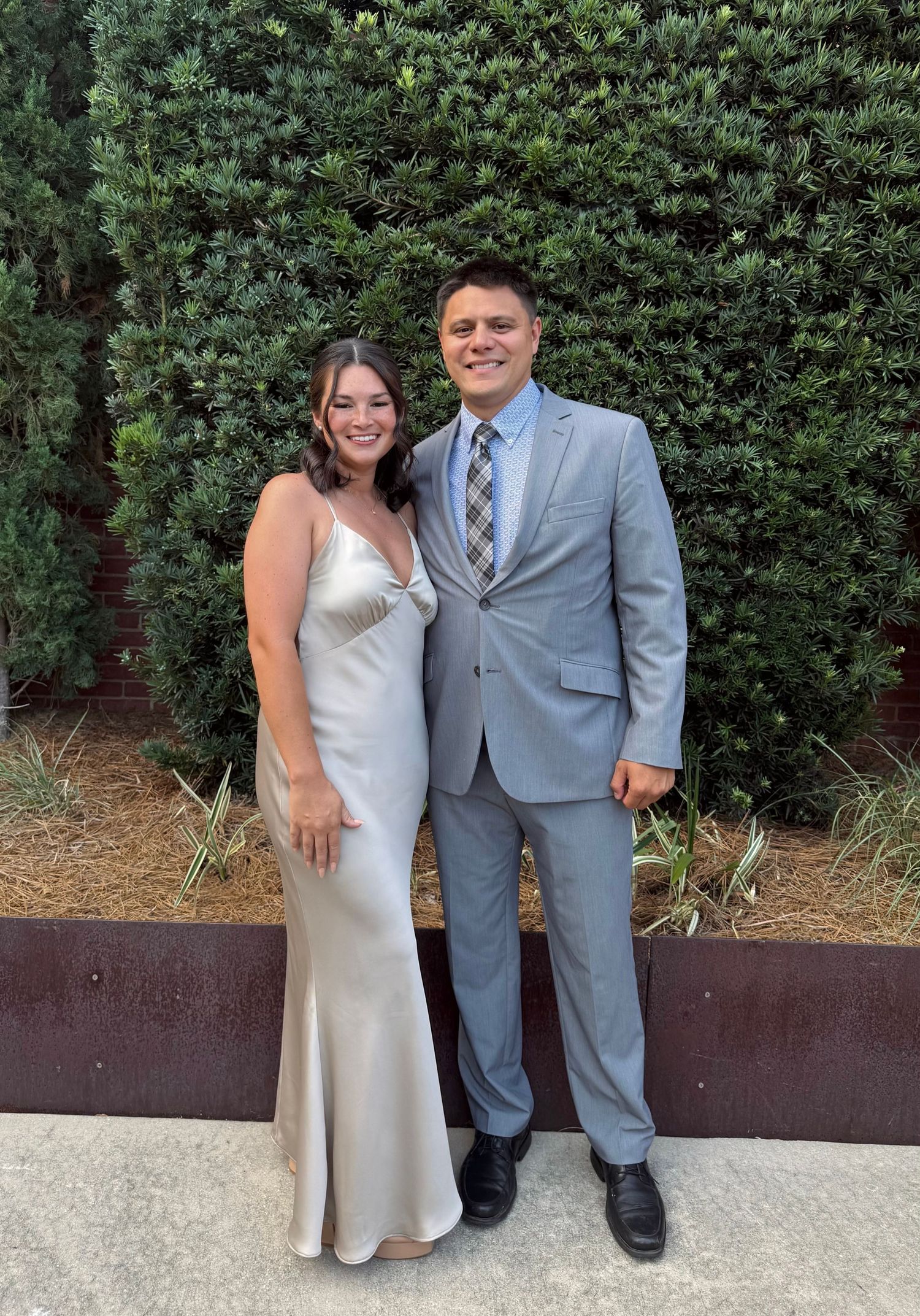 Smiling couple dressed in formal attire posing together in front of a lush green hedge backdrop.