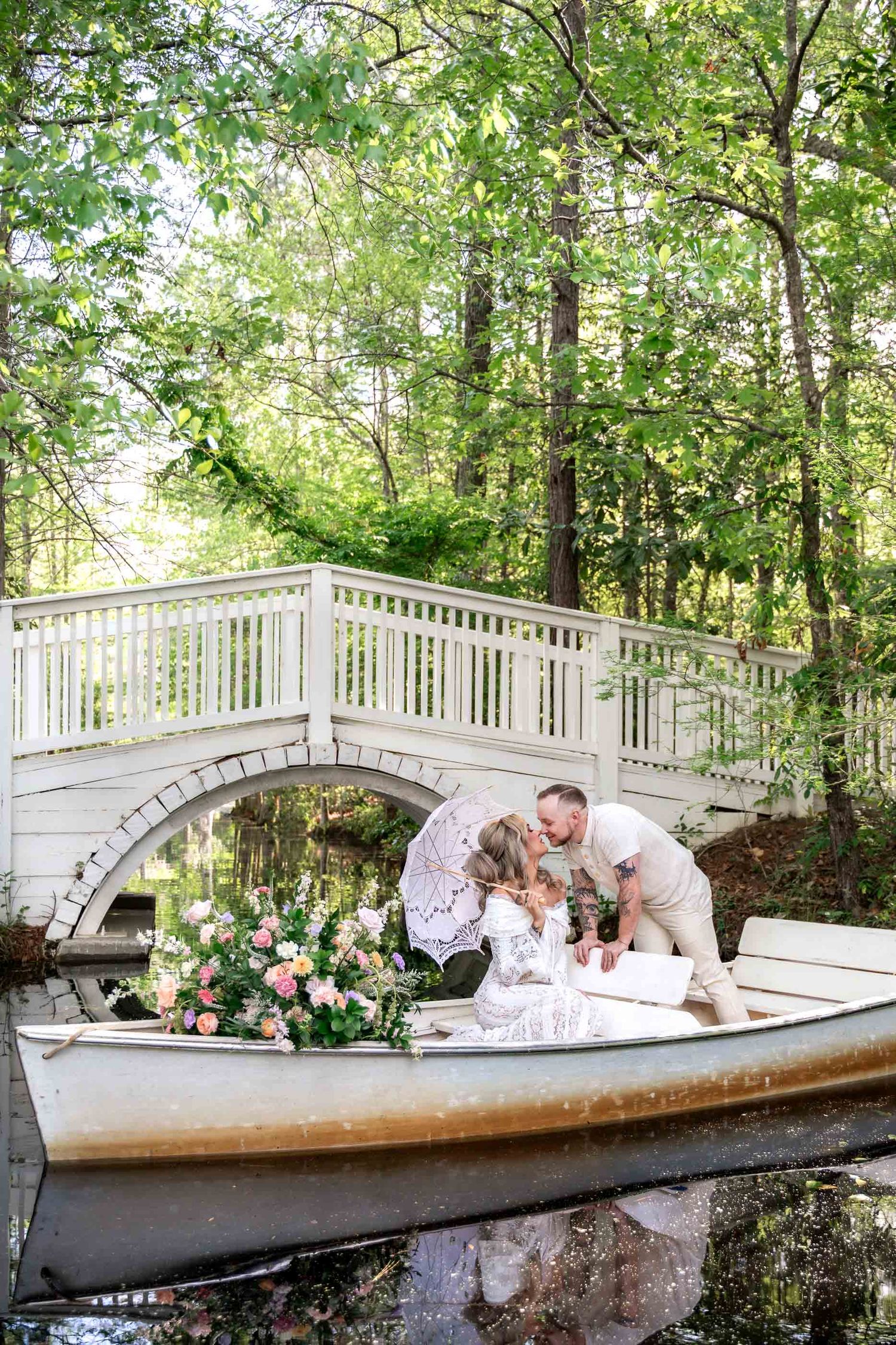 Couple shares a kiss in a decorated rowboat under a white bridge surrounded by lush greenery and pink floral arrangements.