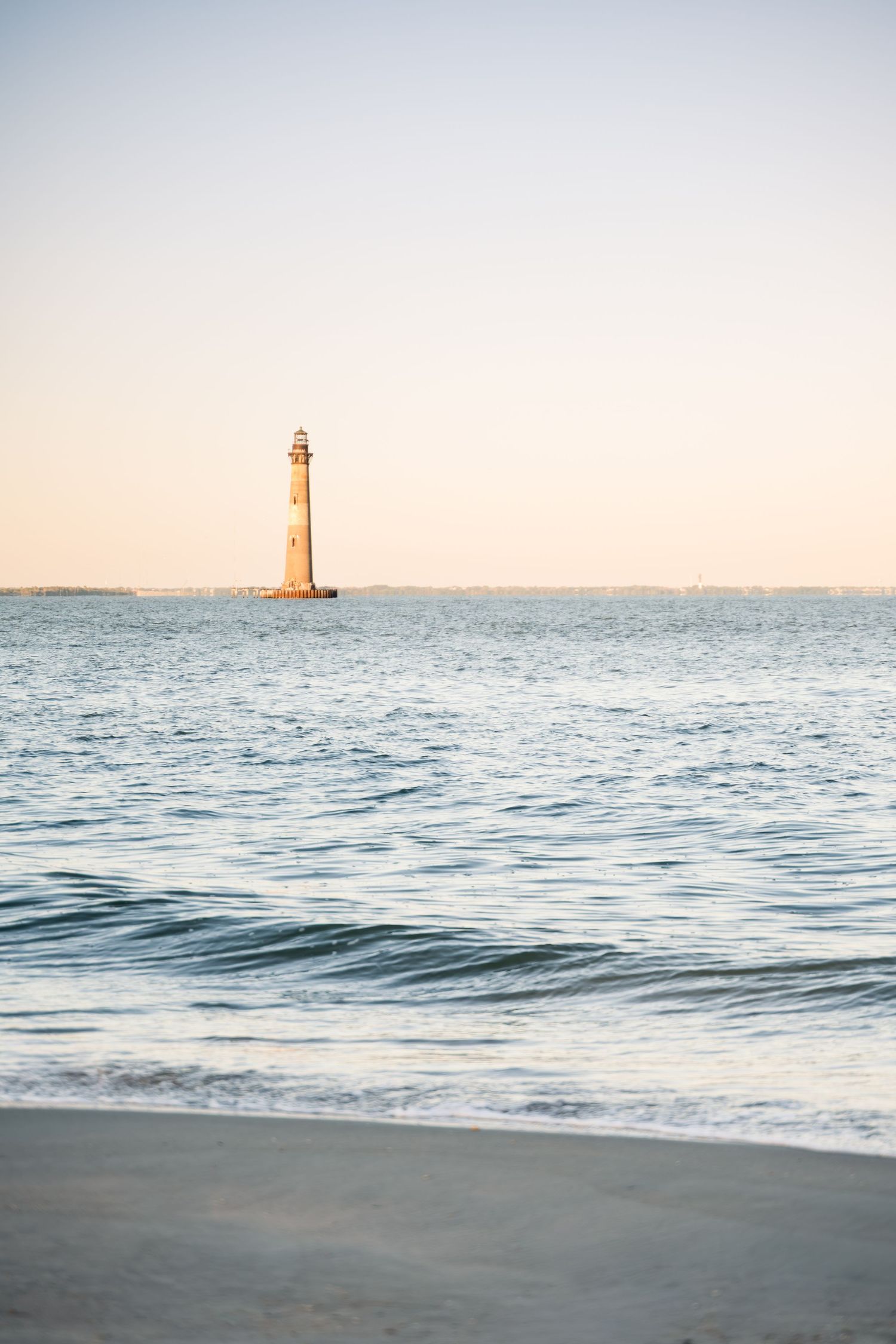 A solitary lighthouse stands in calm ocean waters at golden hour, viewed from a sandy beach shore.