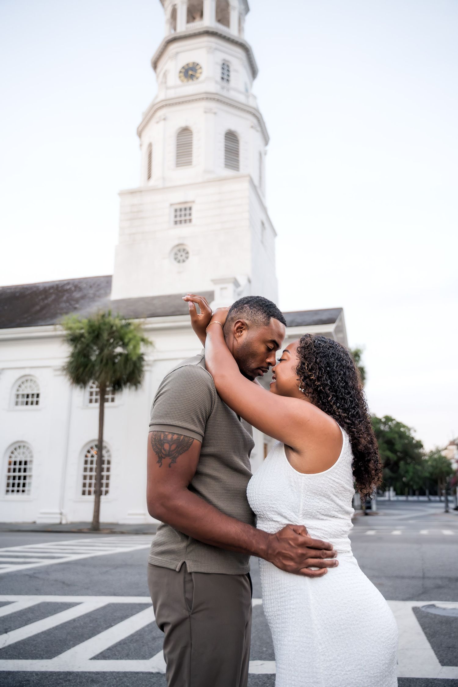 Couple embracing and sharing a romantic moment in front of a white church tower in Charleston, SC.