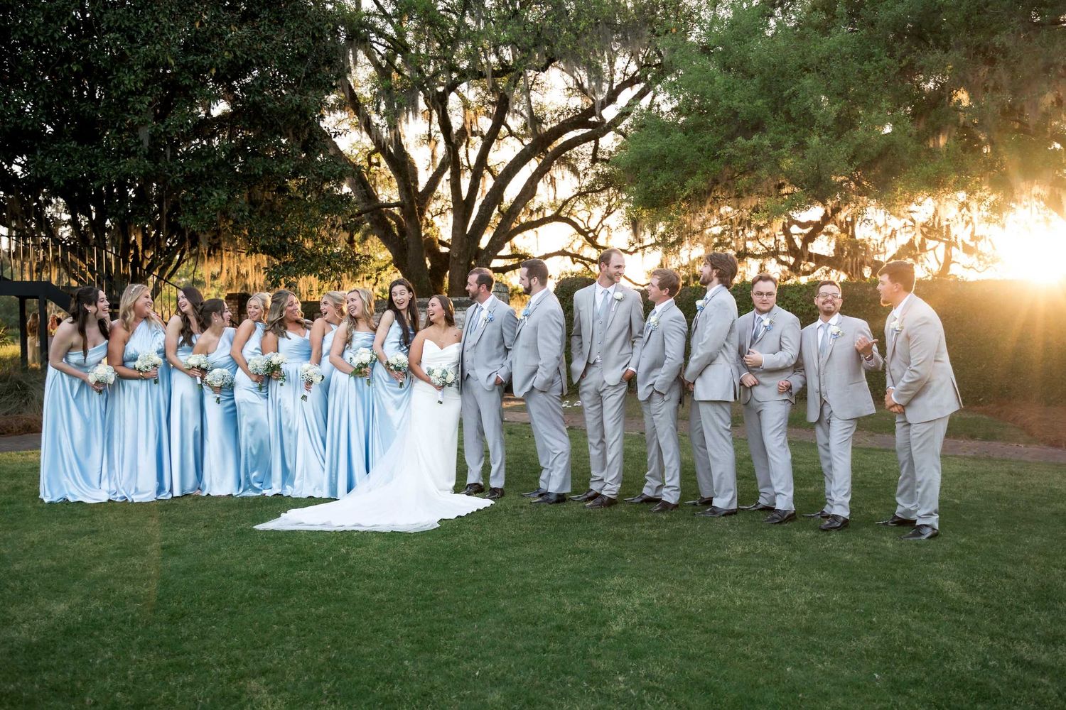 Dunes West Golf and River CluWedding party with bridesmaids in light blue gowns and groomsmen in gray suits posing at sunset under oak tree.