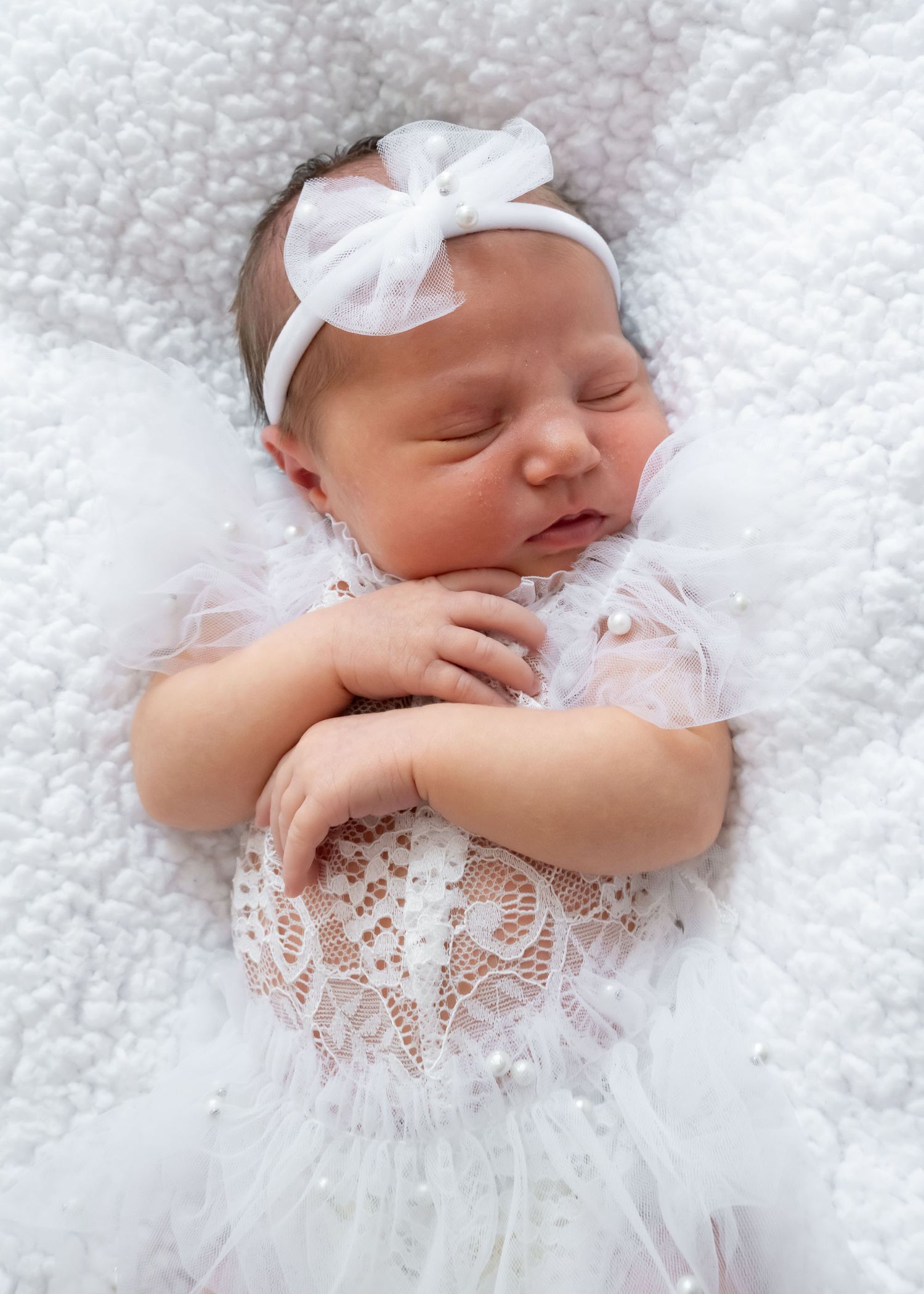 A peaceful newborn baby girl in a white lace dress and bow headband sleeps on a soft white blanket.