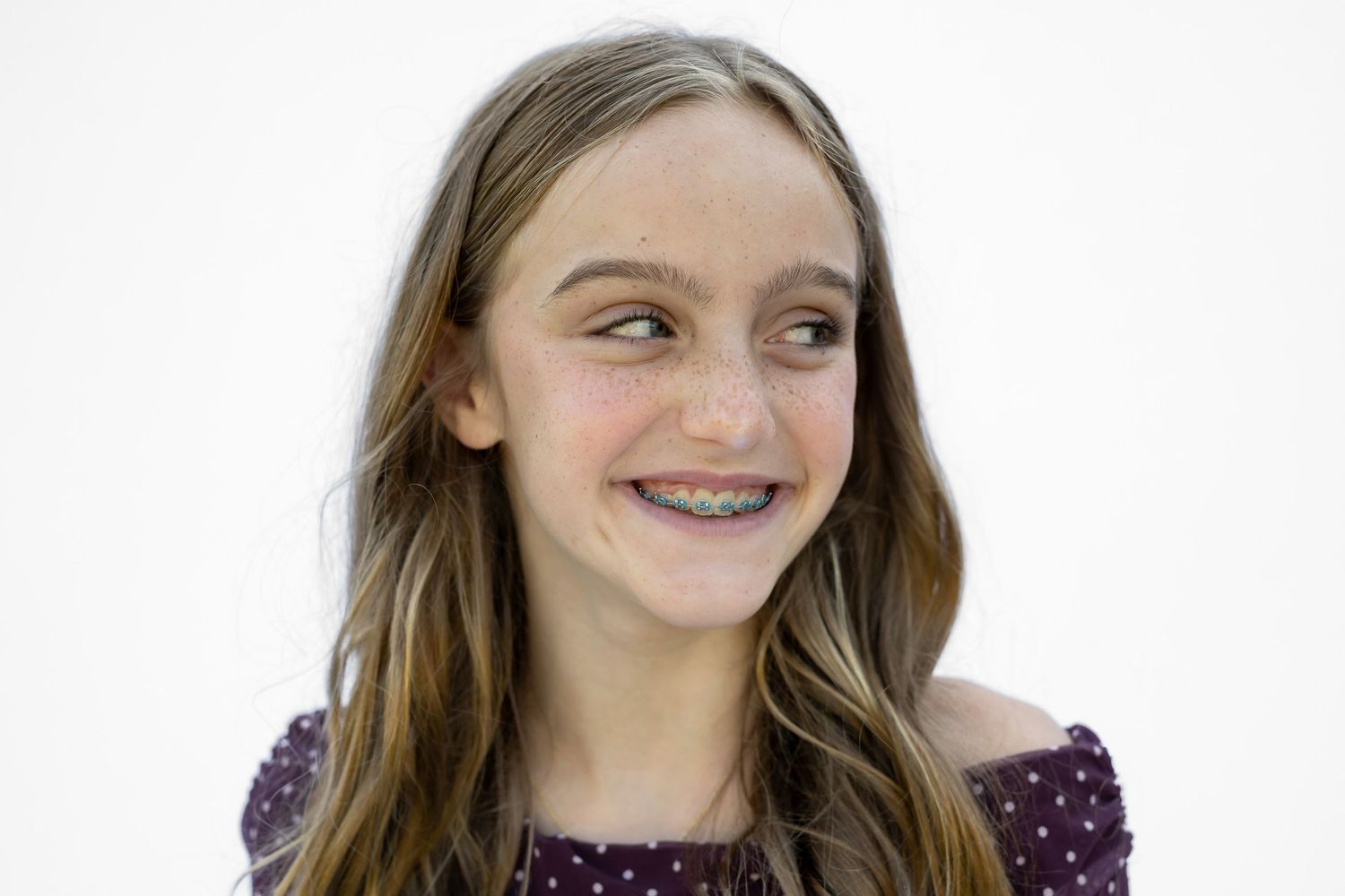 Smiling young girl with braces and long brown hair wearing a purple polka dot top against white background.