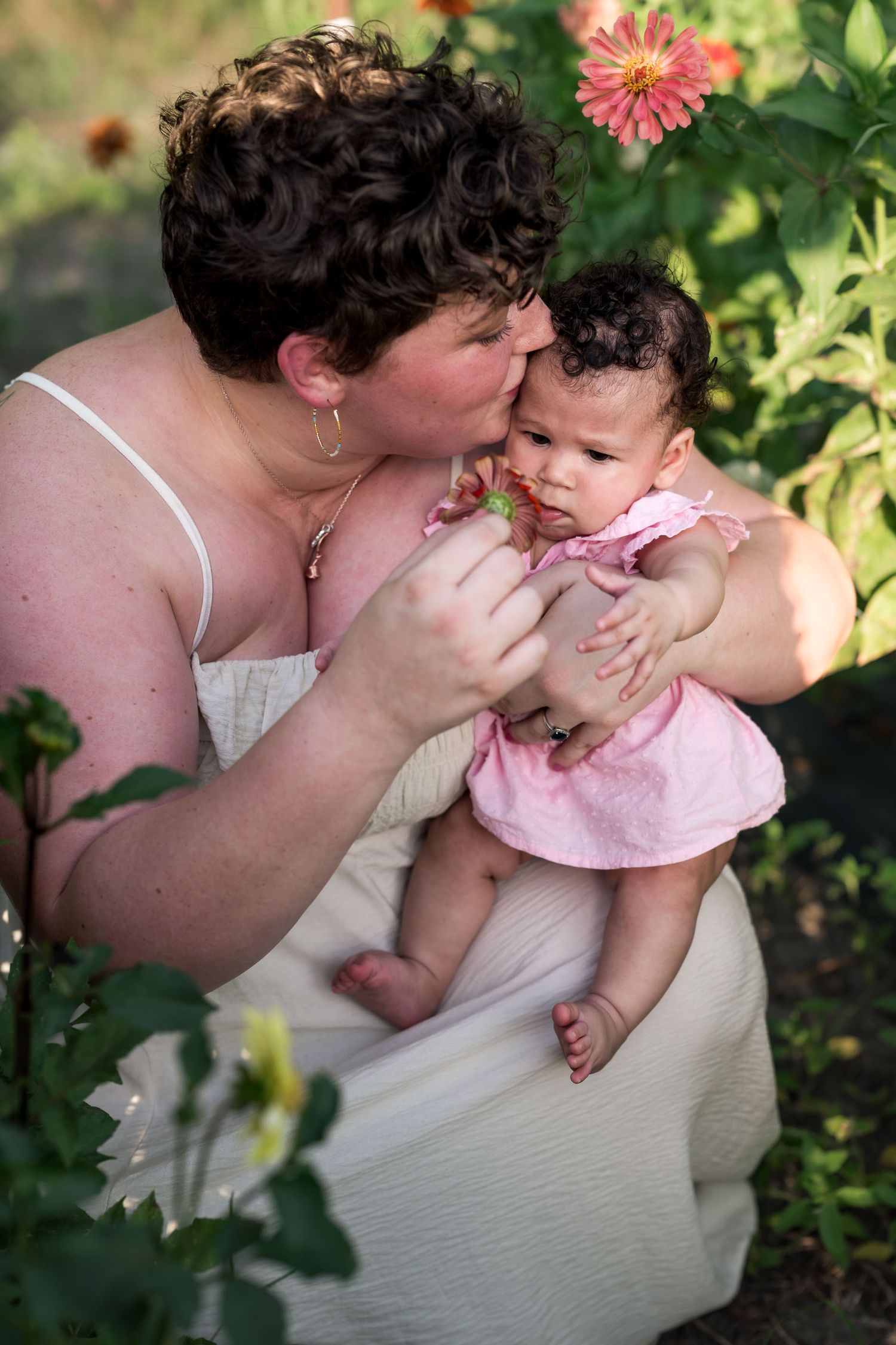 A woman kisses a baby girl in a pink dress while sitting among colorful garden flowers.