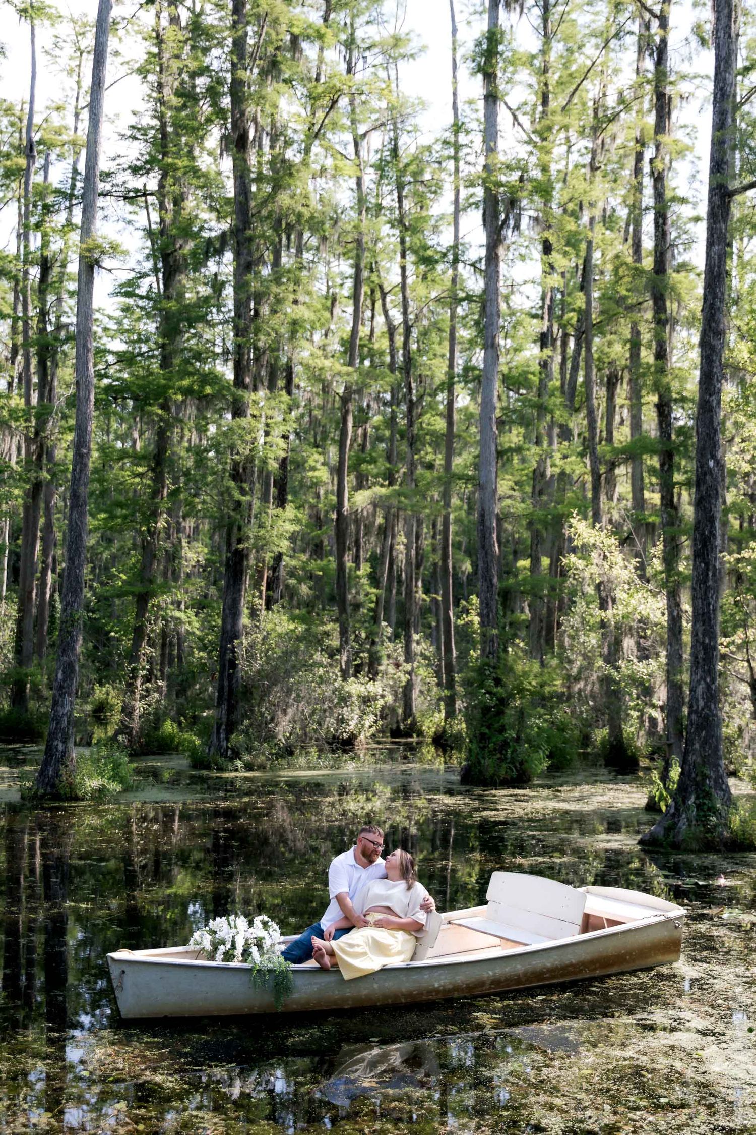 Couple sitting in a white rowboat surrounded by cypress trees in a lush green swamp, with white floral arrangements.