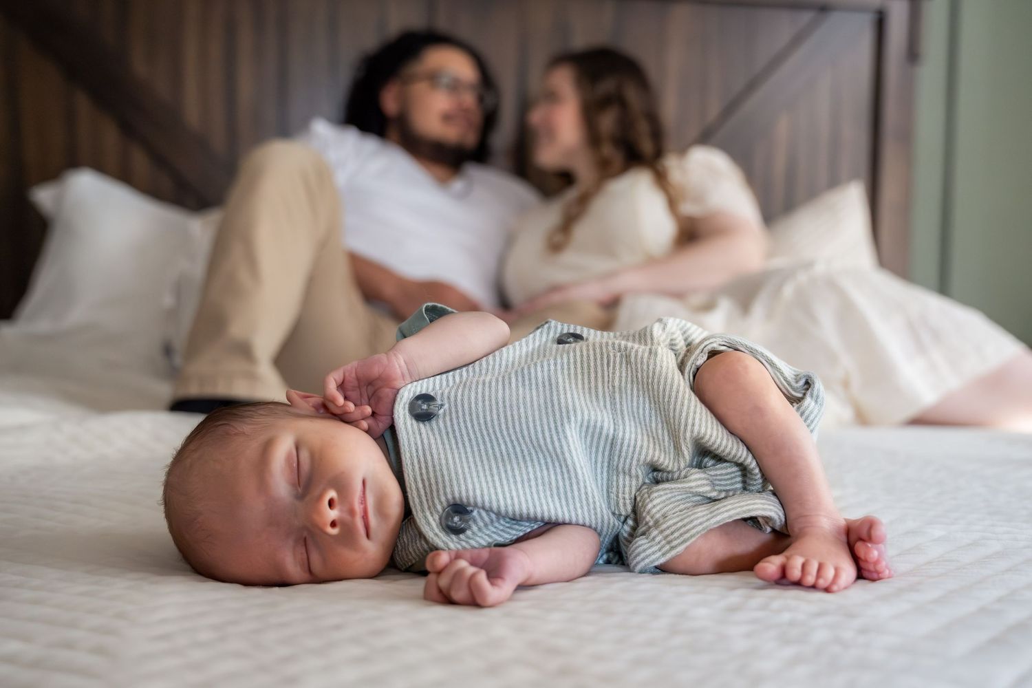 New parents relax on bed while their newborn baby sleeps in striped outfit in the foreground.