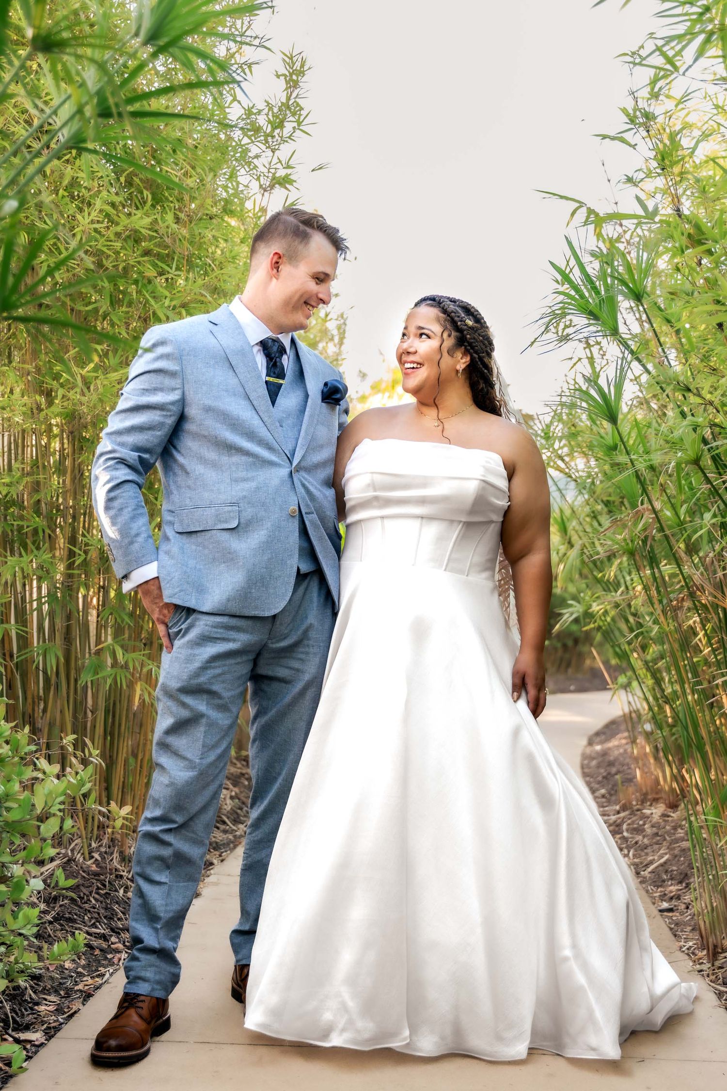 Bride in white strapless gown and groom in light blue suit walking together through lush bamboo garden path.