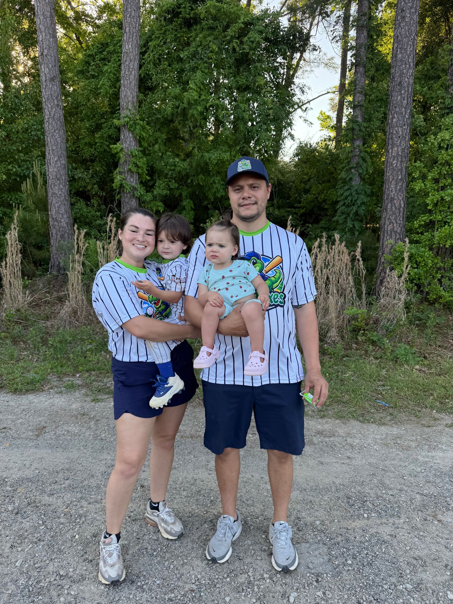 Family of four wearing matching baseball jerseys posing together outdoors with two young children.