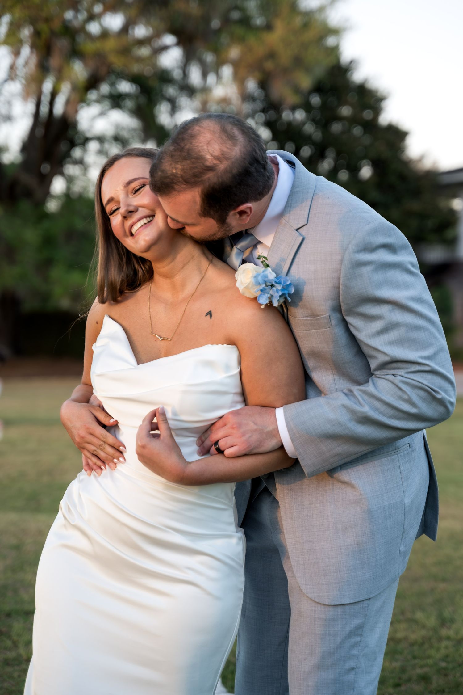 Groom in gray suit kissing bride on cheek as she smiles joyfully in white strapless wedding dress outdoors.