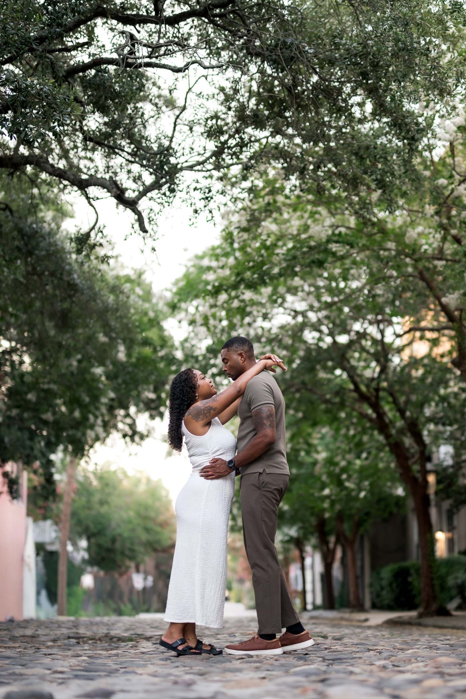A romantic couple embraces on a cobblestone street surrounded by lush green trees in a charming Southern city.