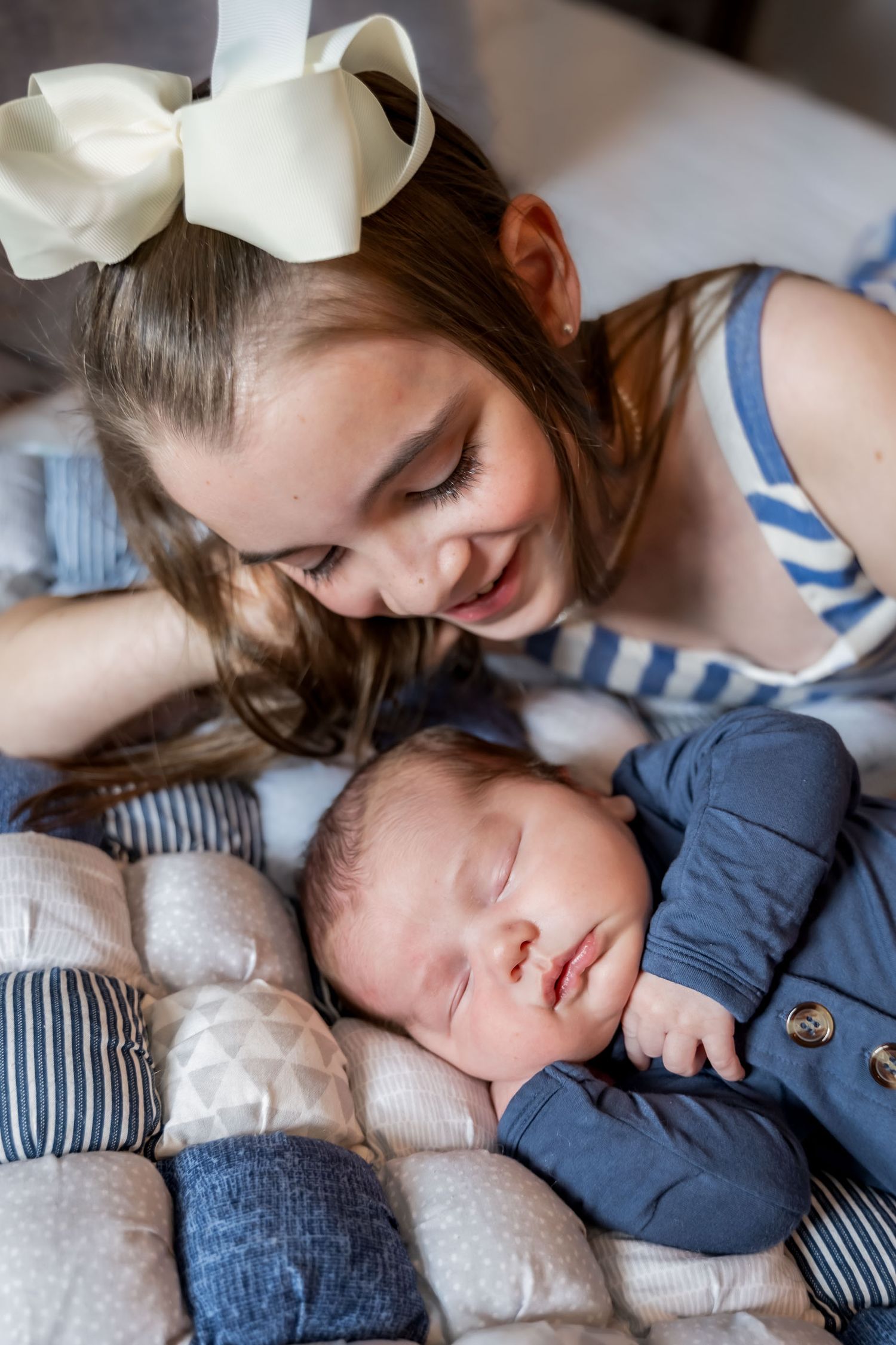 Young girl with white bow smiles at sleeping newborn baby in blue onesie on patchwork pillow.