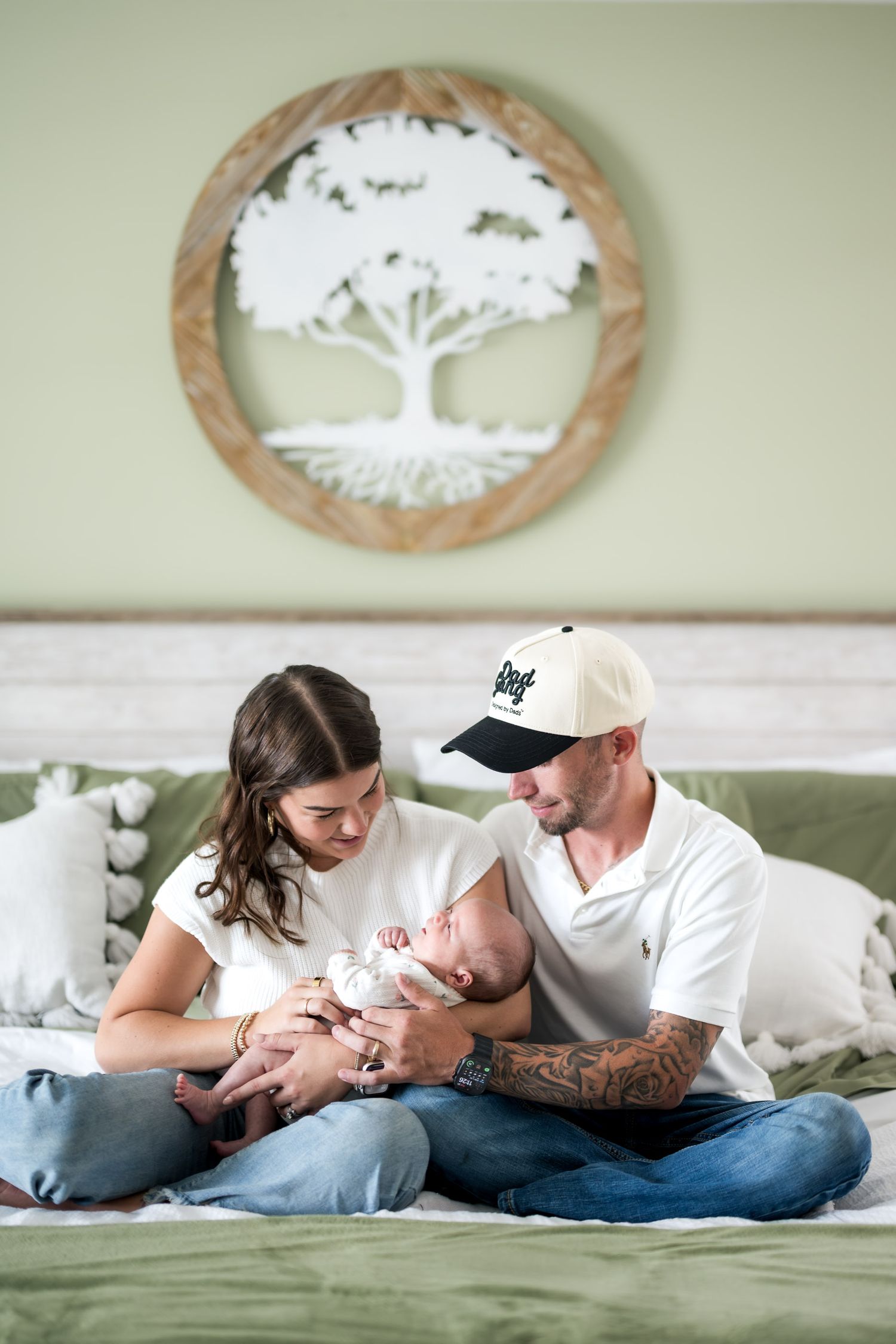 New parents sitting on a bed holding their newborn baby beneath a decorative tree of life wall art.