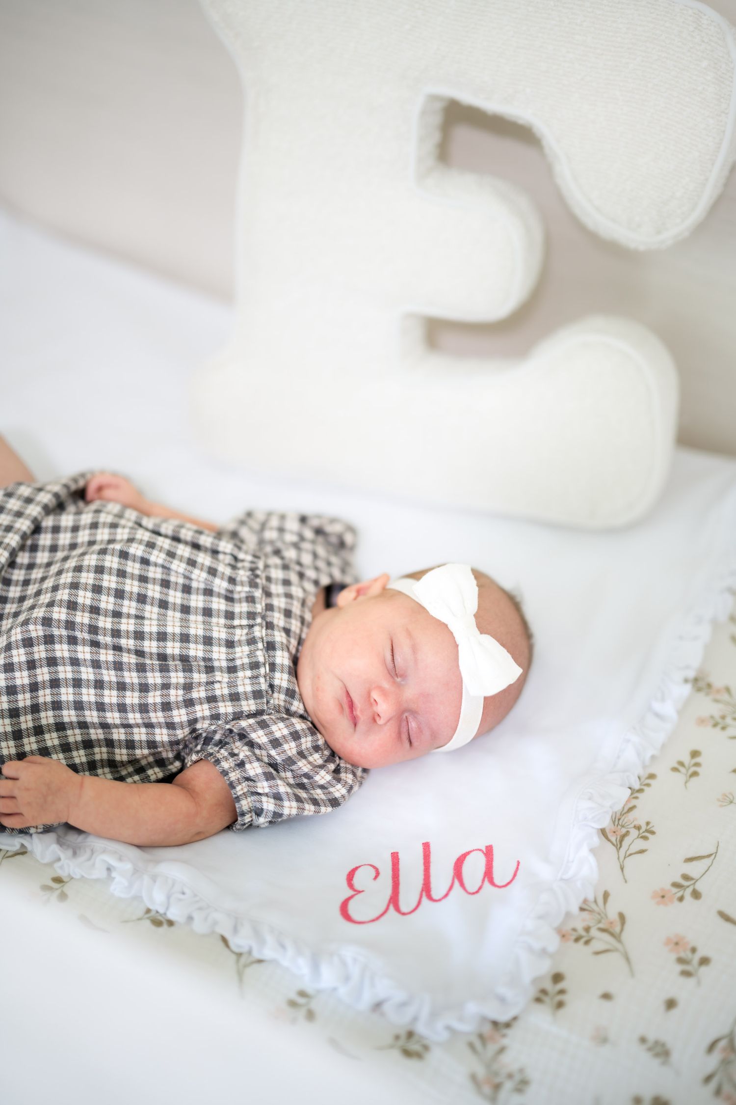 Newborn baby girl named Ella sleeping peacefully on a floral blanket wearing a plaid outfit and white bow headband.