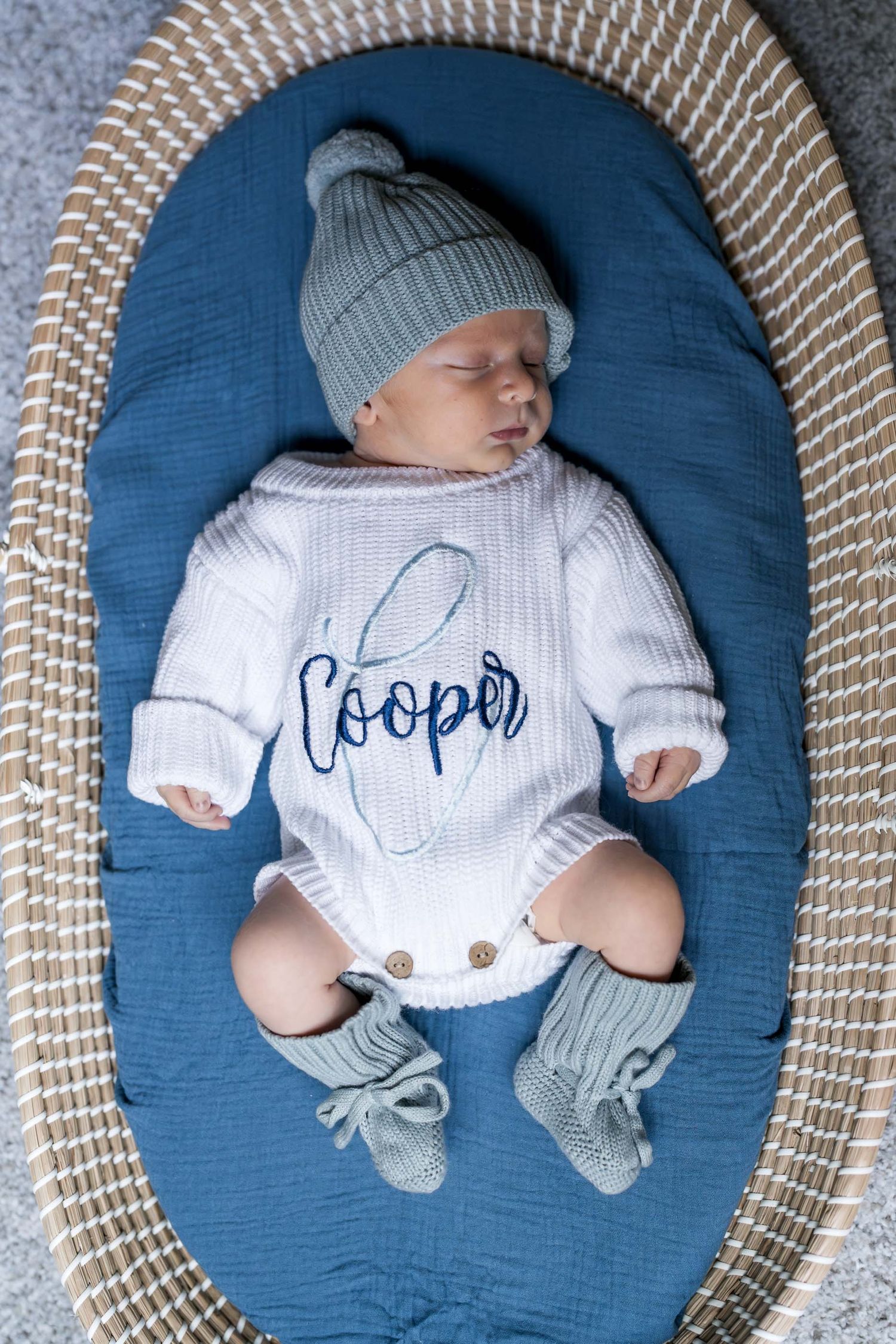 Newborn baby named Cooper sleeping in a blue oval nest, wearing a white knit romper and sage green beanie with booties.