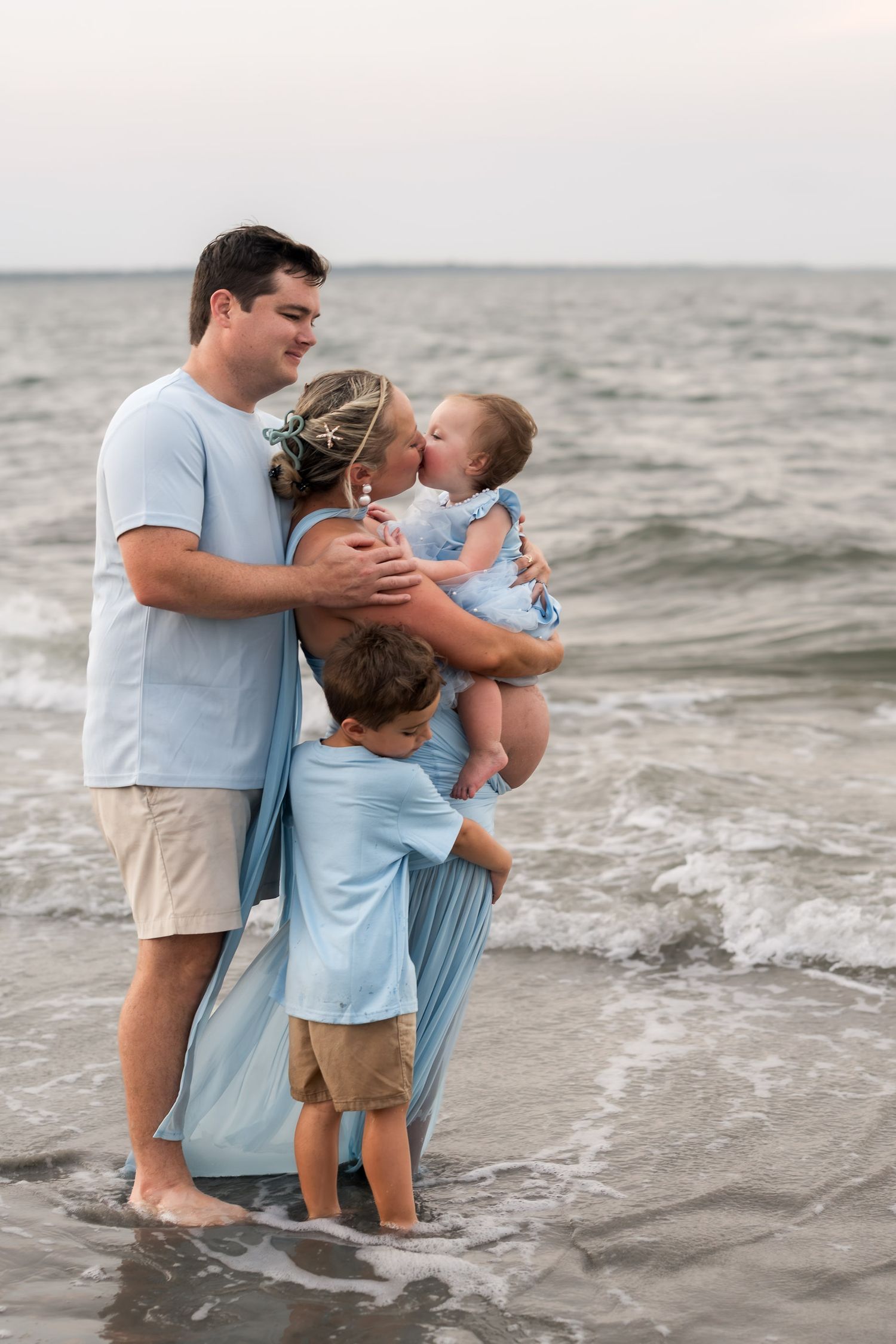 Family of four in light blue outfits standing together on a beach shoreline with ocean waves behind them.