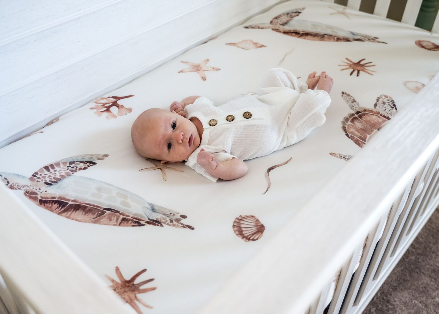 Peaceful newborn baby in white onesie lying in crib with ocean-themed seashell bedding.