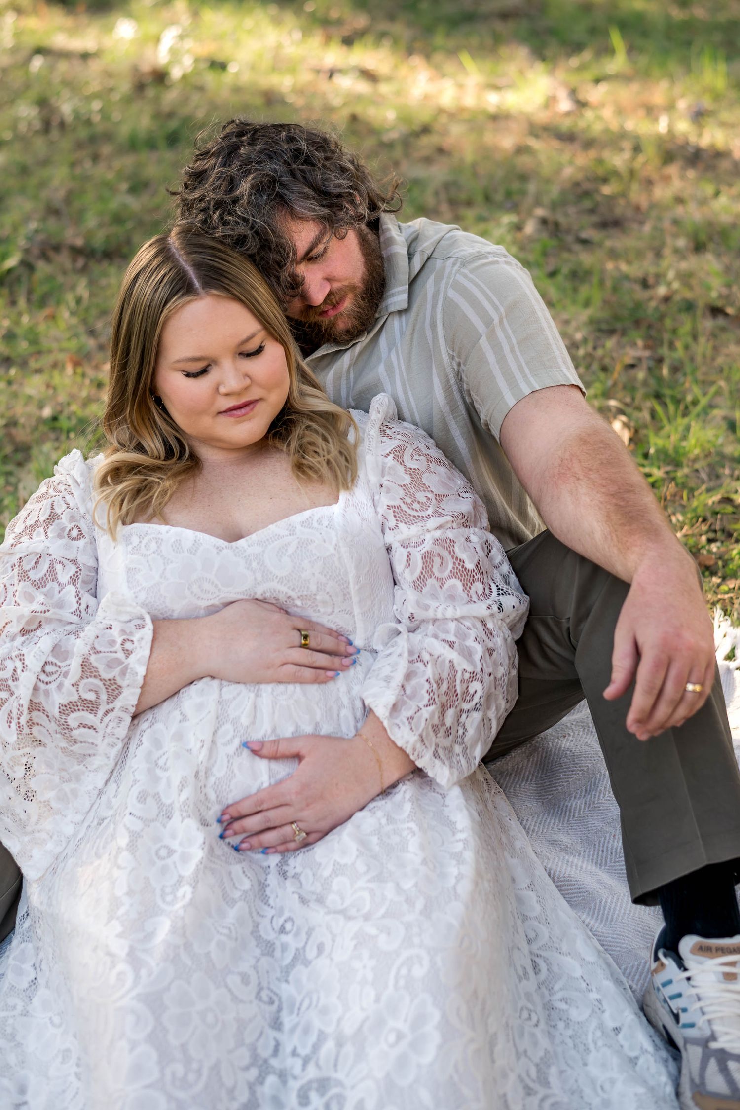 Moncks Corner.Pregnant woman in white lace dress sitting with partner outdoors in soft sunlight.
