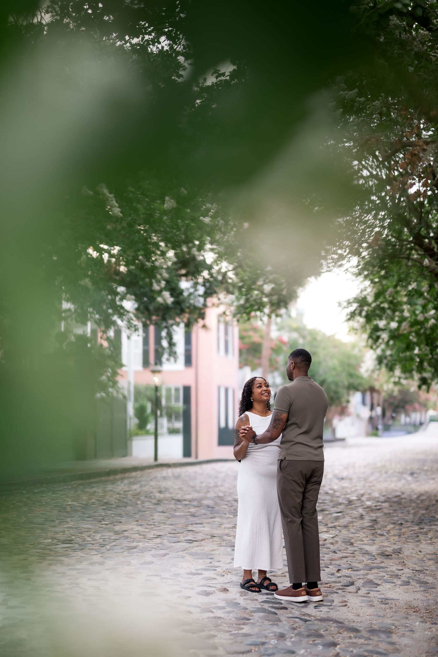 Couple sharing a tender moment on a cobblestone street framed by lush green trees in Charleston.