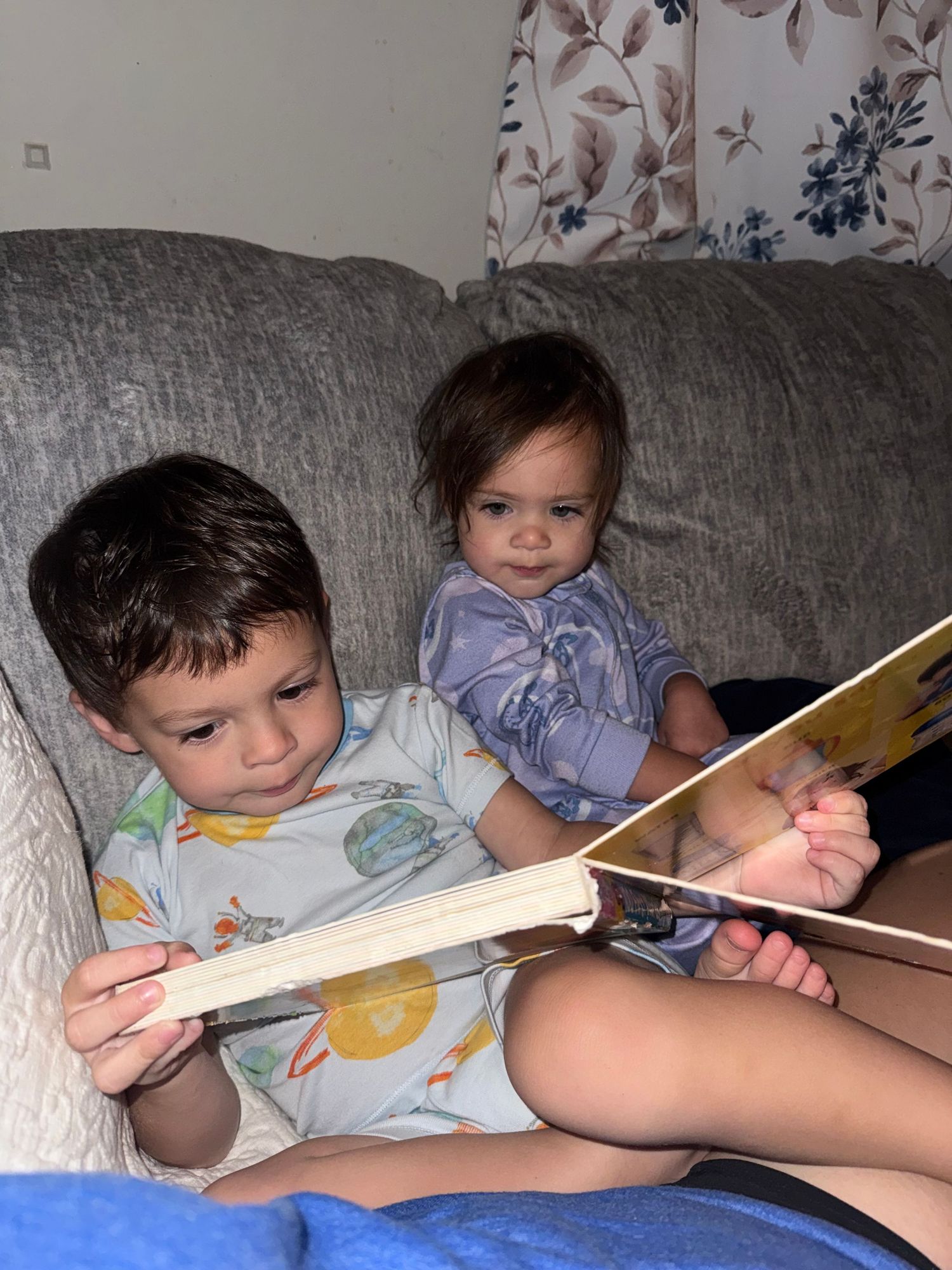 Two young children sitting on a gray couch, with an older toddler reading a picture book to a smiling baby.