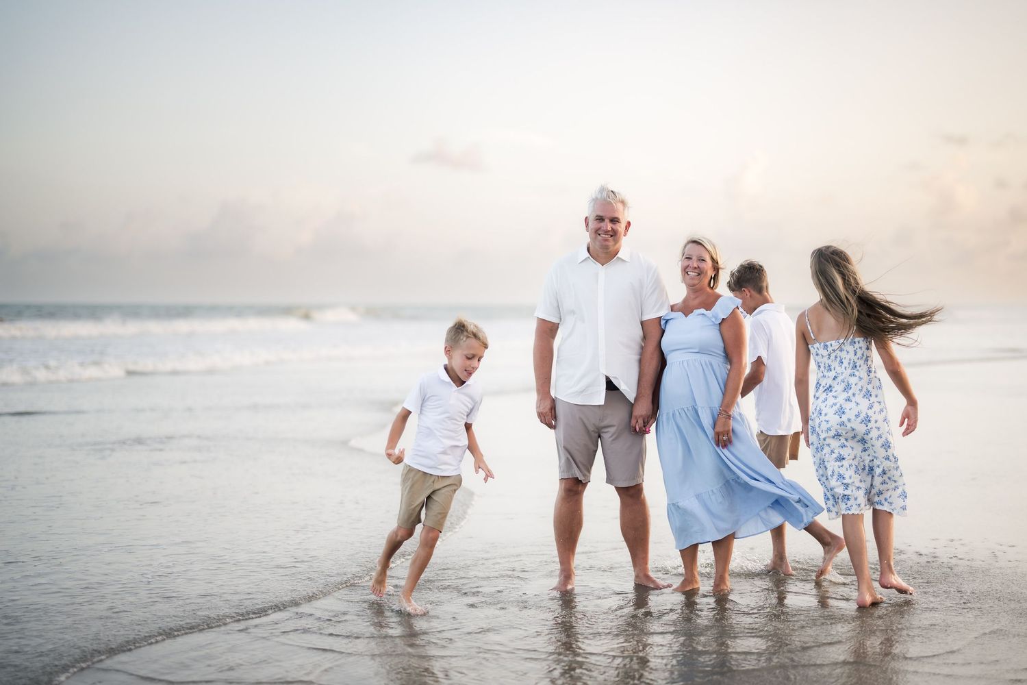 Family of five enjoying a beach sunset, with grandparents and children playing in shallow ocean waves on a sandy shore.