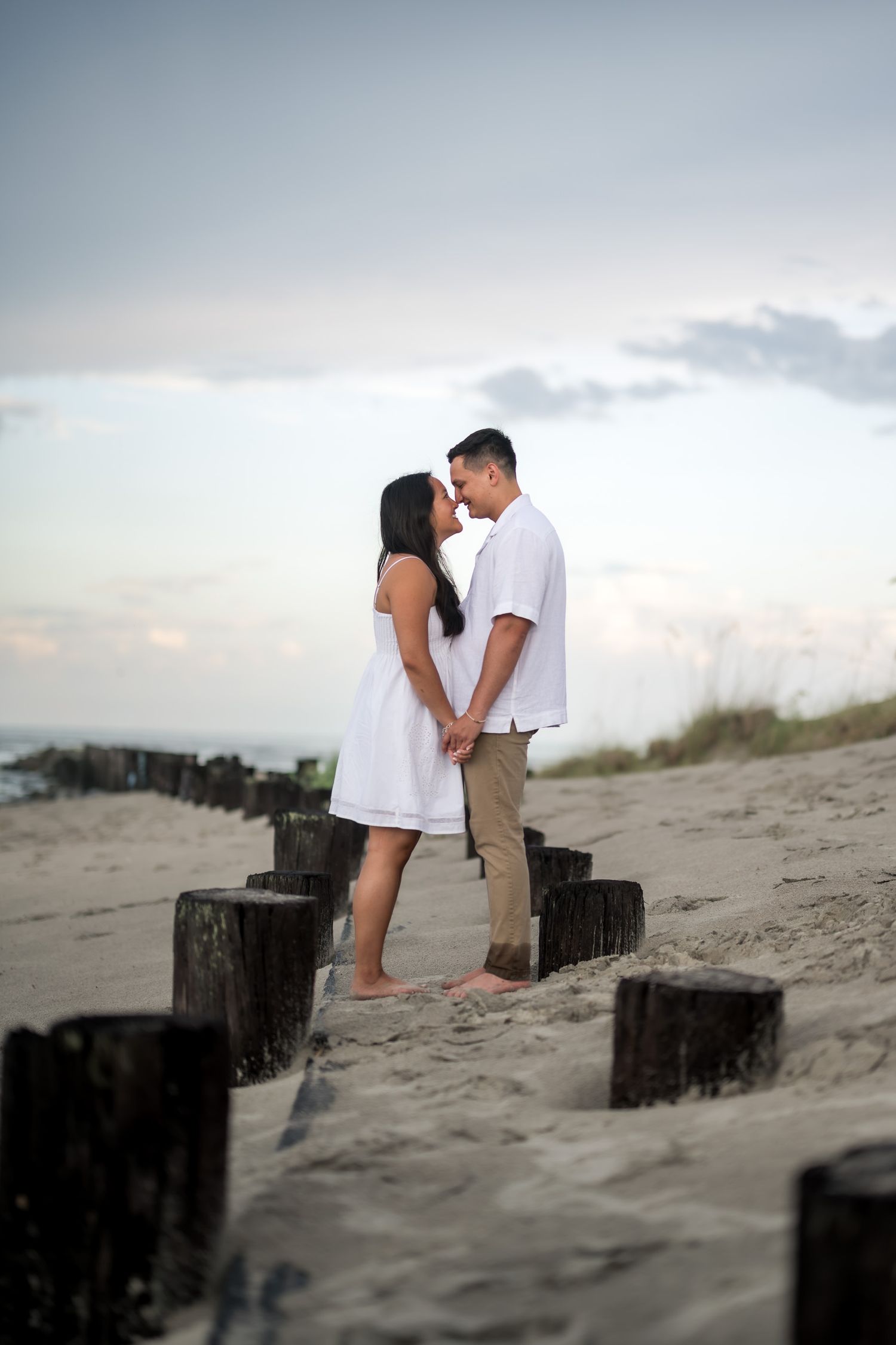 Couple kissing on sandy beach at sunset, holding hands between rustic wooden posts, wearing white outfits.