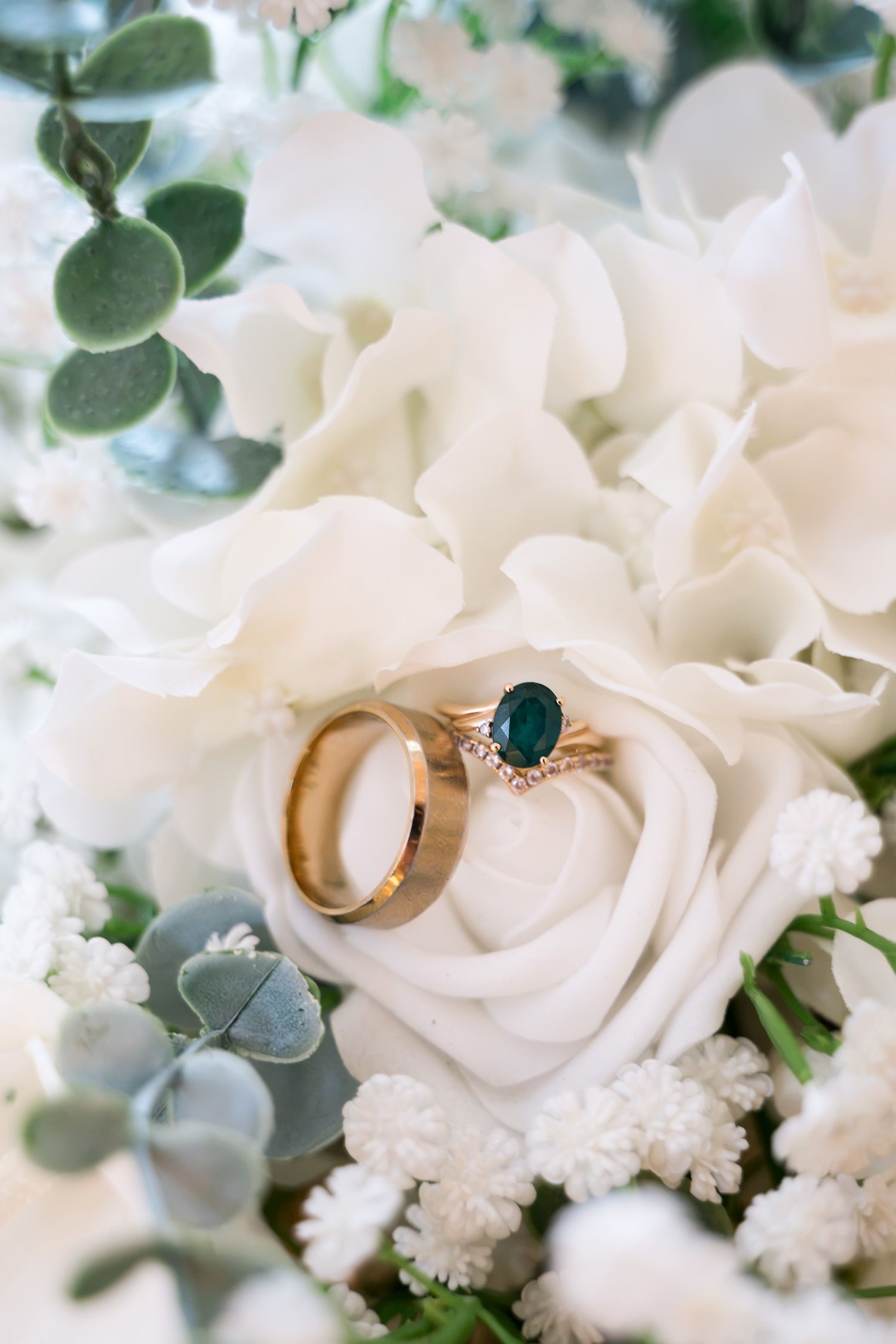 Gold wedding bands and emerald engagement ring displayed on white roses and eucalyptus bridal bouquet.