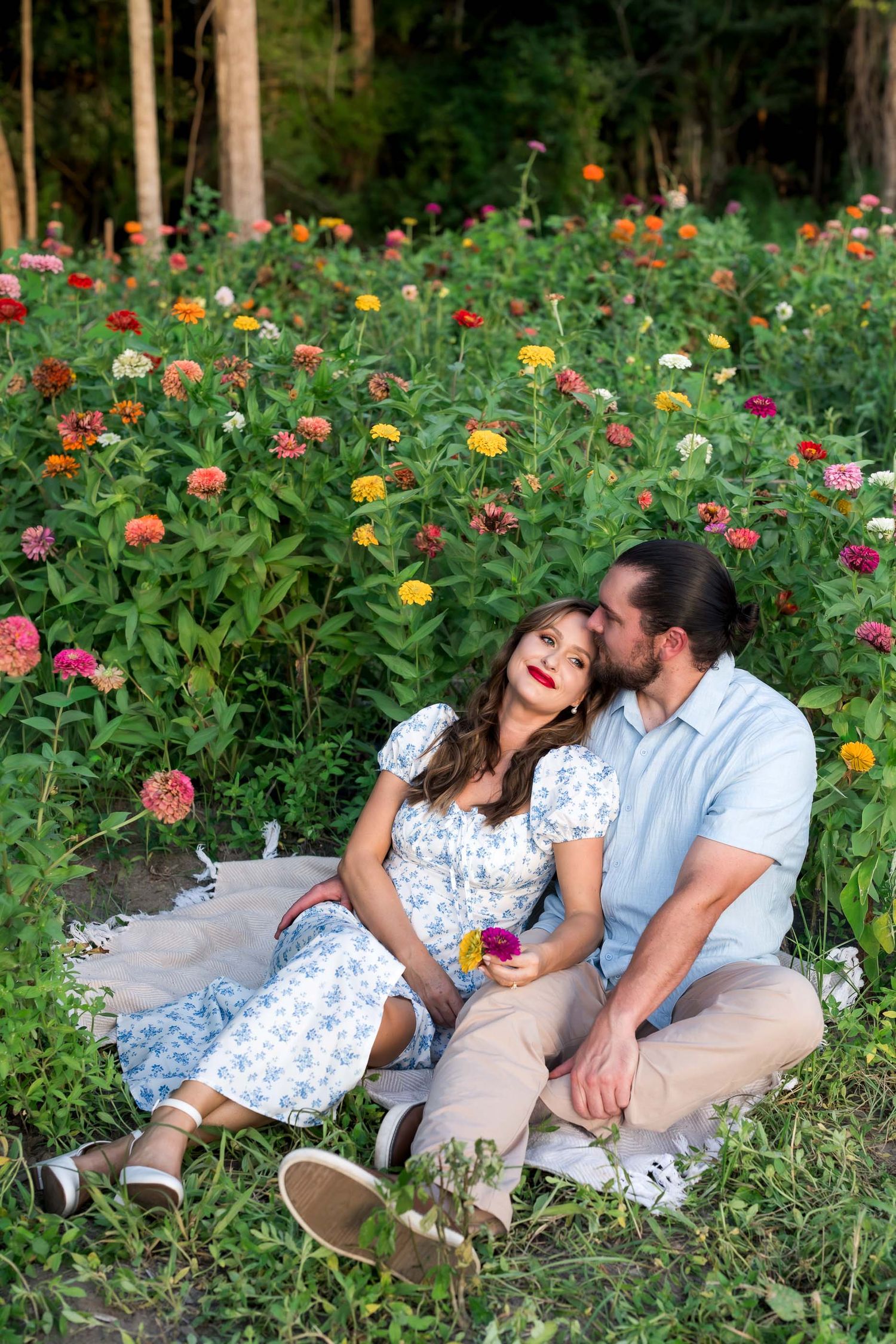 Couple sitting in a colorful flower garden, woman leaning back as man kisses her cheek, wearing casual summer outfits.