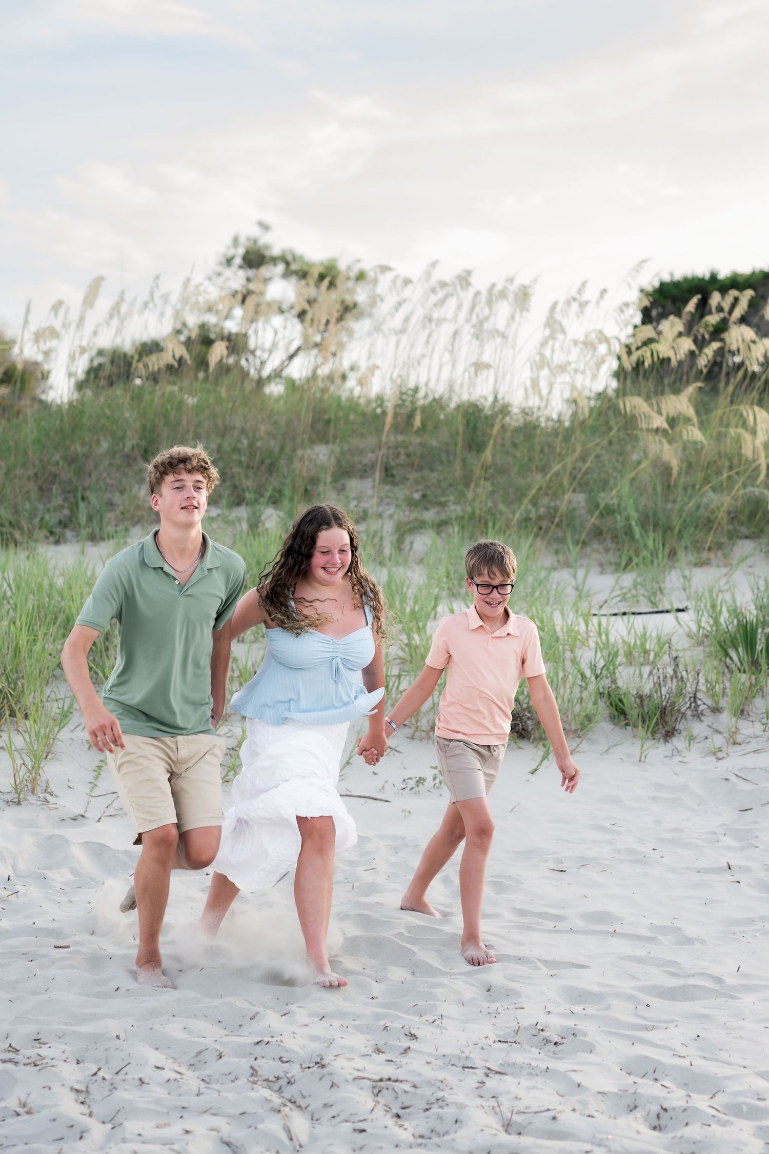 Three children laughing and running barefoot on sandy beach dunes with sea oats in background.