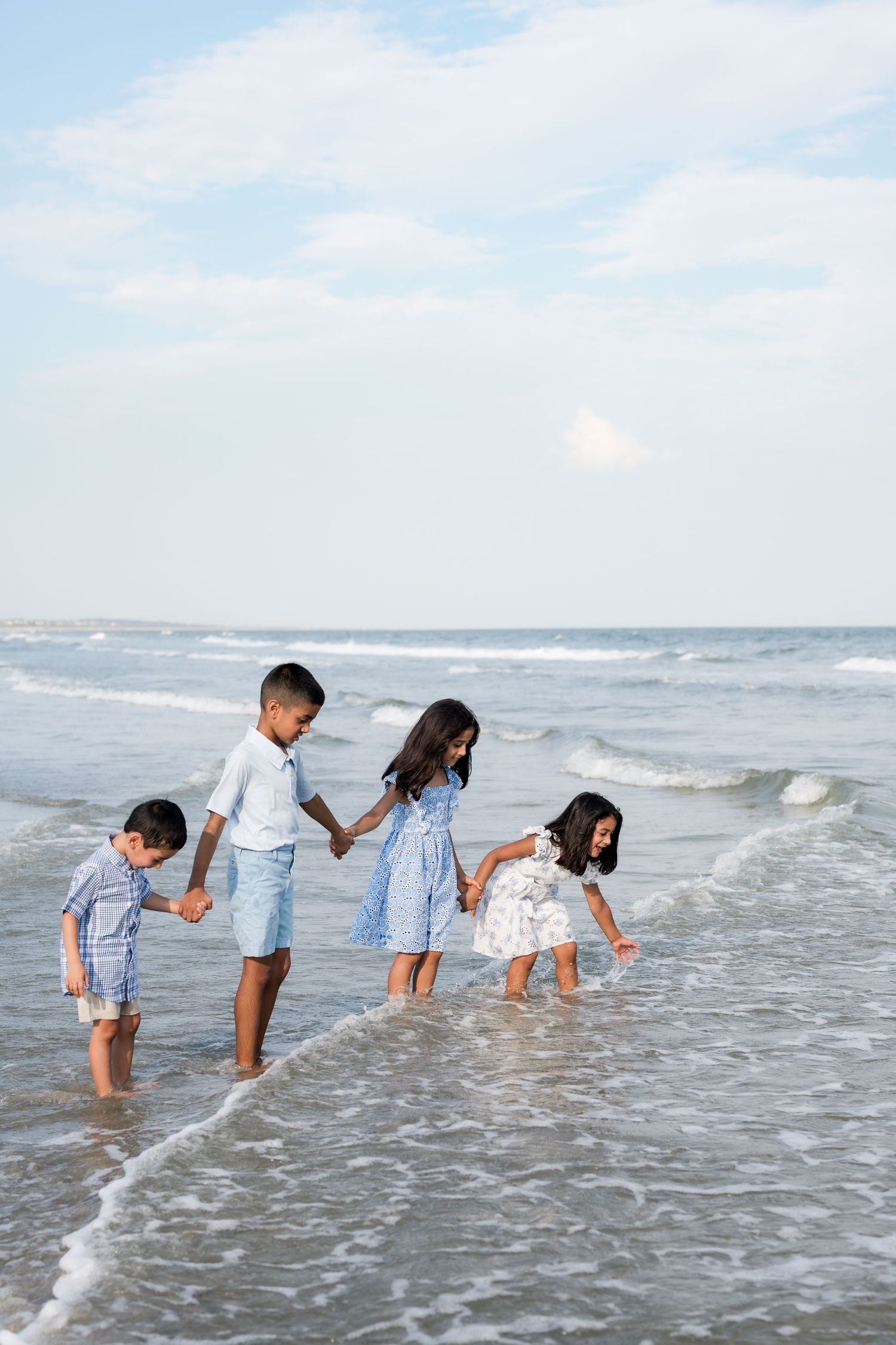 Four children in blue and white outfits hold hands while playing in ocean waves on a sunny beach.