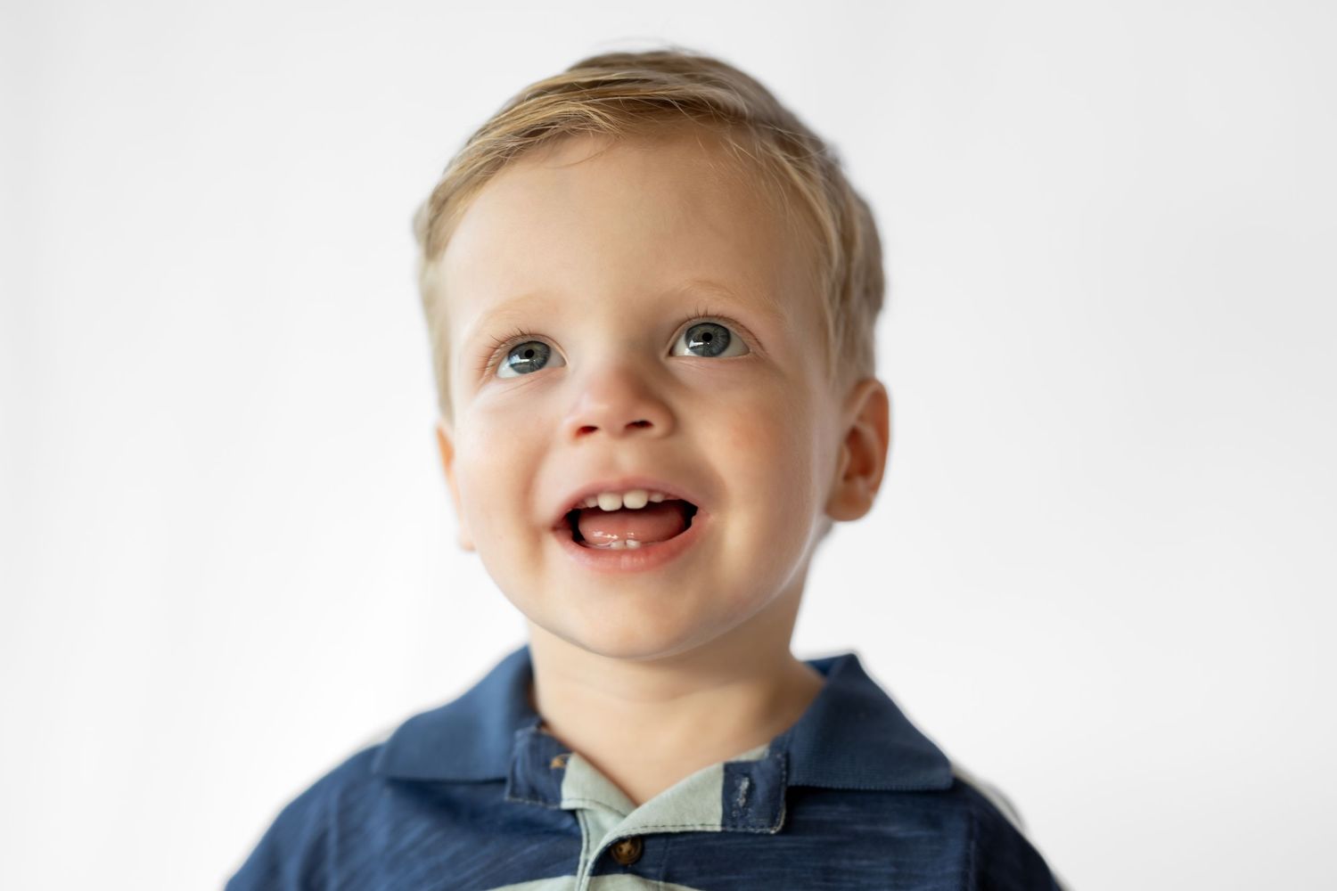 Smiling toddler boy with blue eyes and blonde hair wearing a blue shirt against a white background.