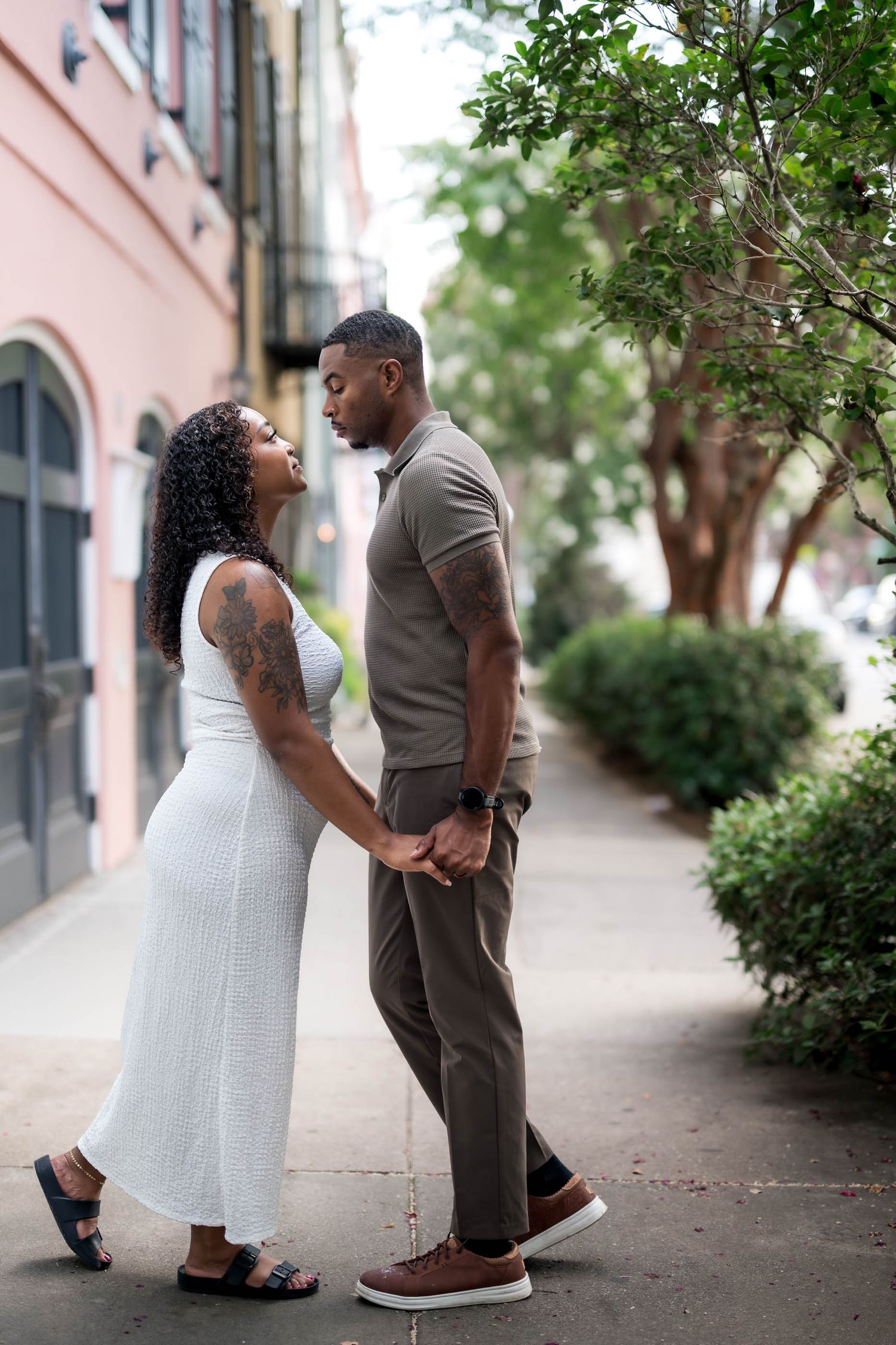 A couple stands face to face holding hands on a sidewalk, woman in white midi dress and man in olive polo shirt.