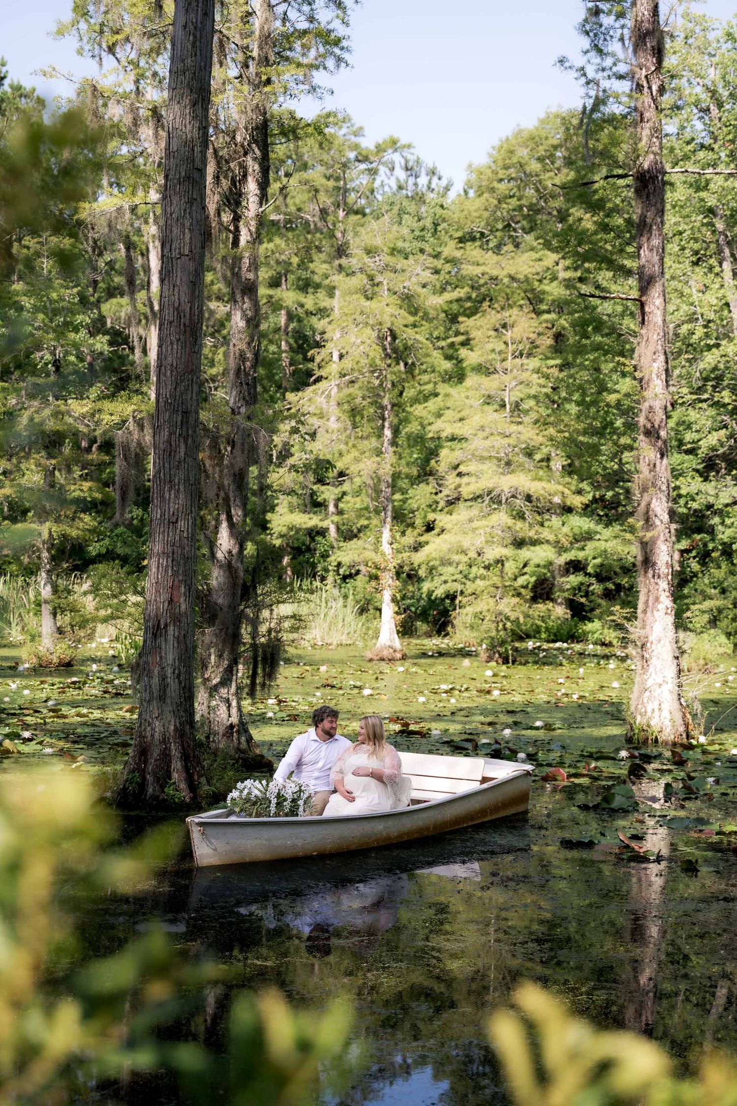 Couple sitting together in a white rowboat surrounded by lily pads and cypress trees in a serene swamp.