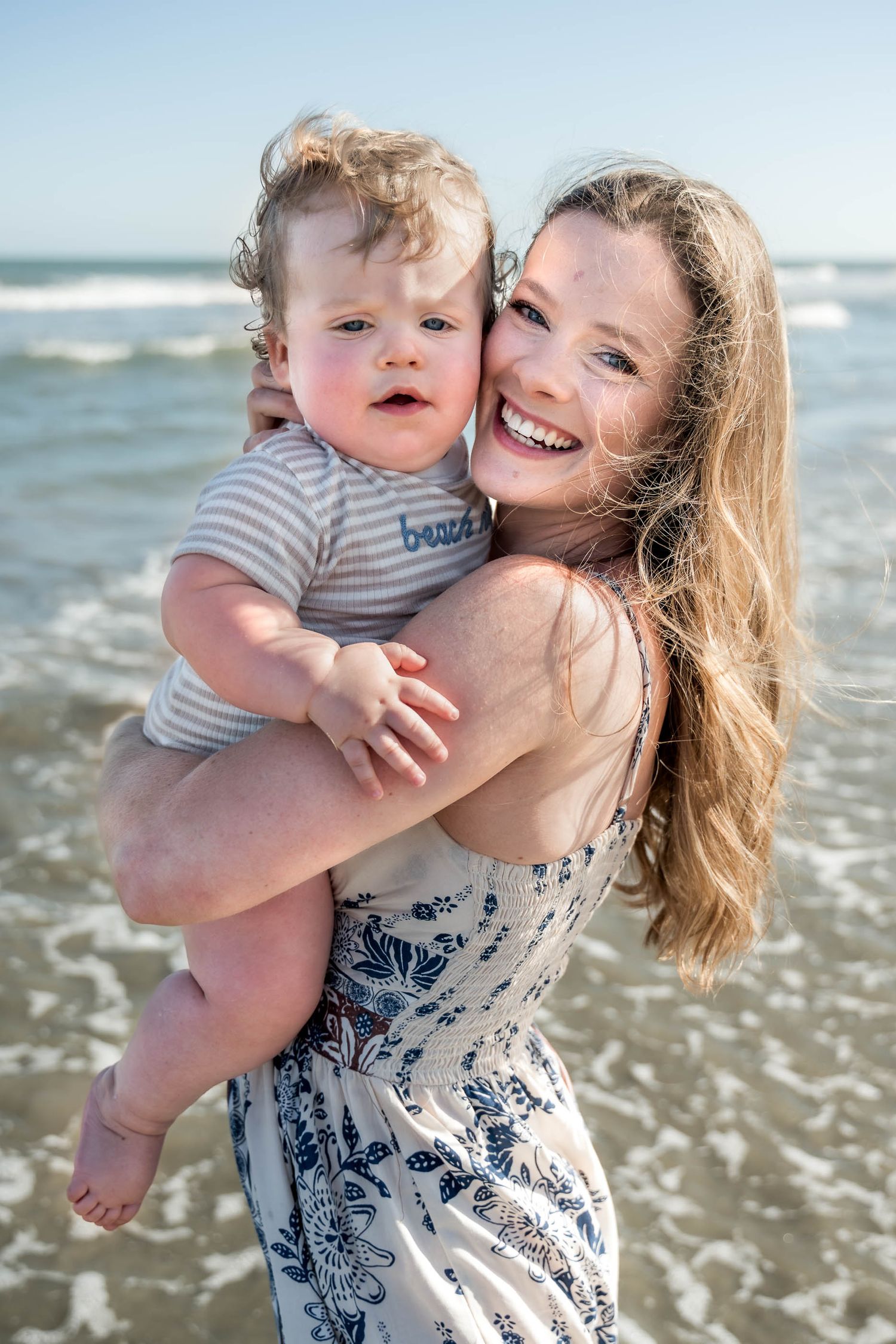 Isle of Palms. A smiling mother with long hair holds her adorable baby boy on a sunny beach with ocean waves.