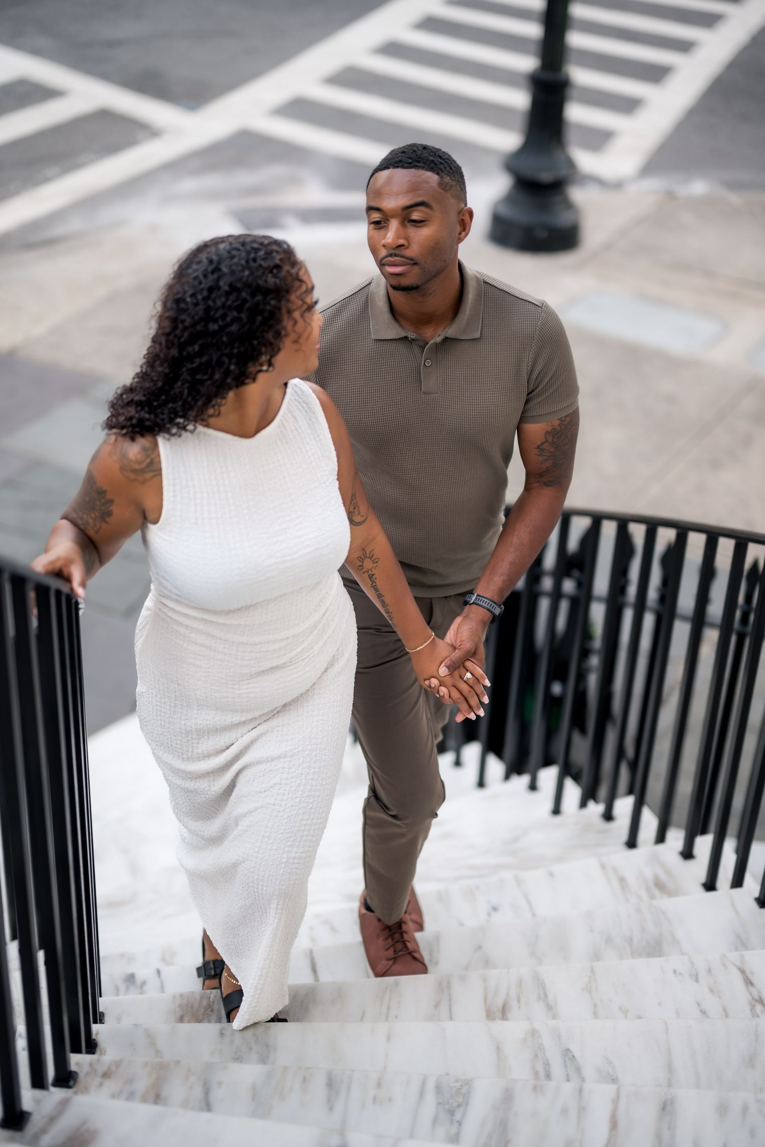 A couple holds hands while walking up marble stairs, dressed in white dress and olive polo shirt.
