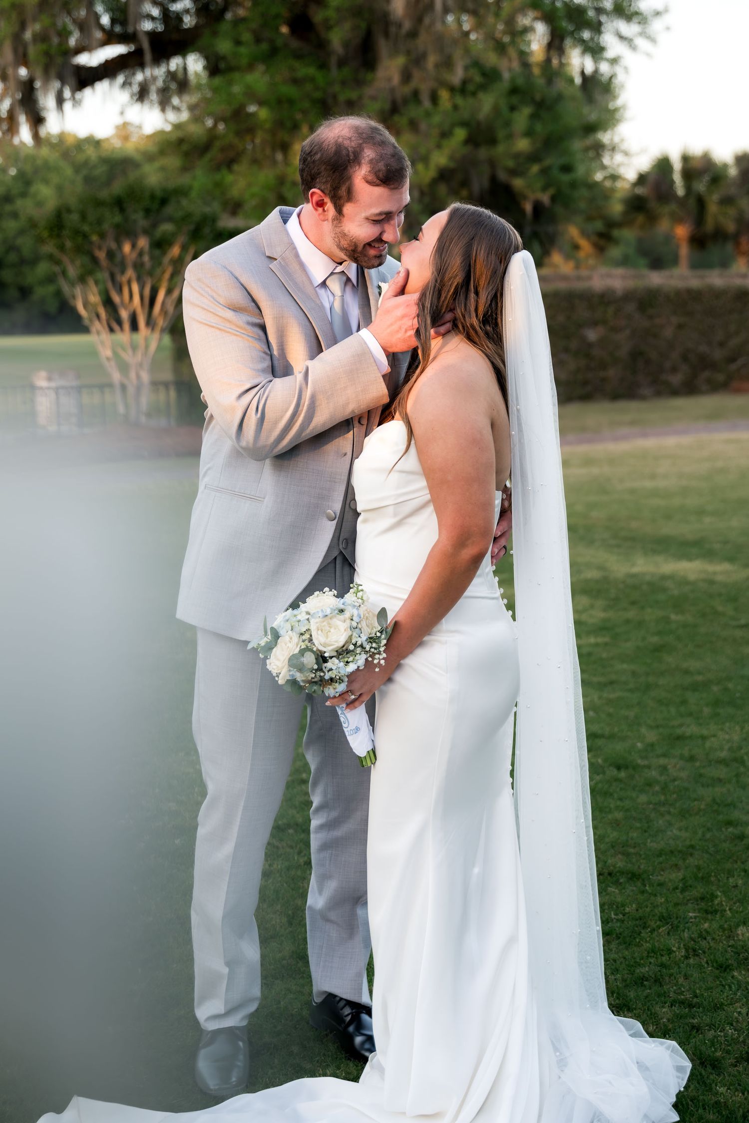Dunes West Golf and River Club. Bride and groom sharing romantic moment on green lawn, bride in white gown with veil holding white bouquet.